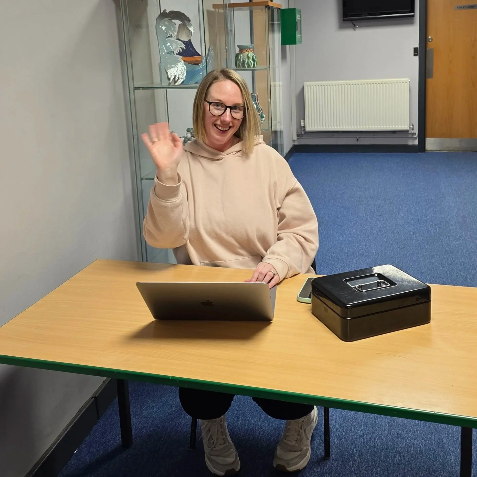 A woman with glasses waving while sitting at a table with a laptop, a black cash box, and a smartphone in a room with blue carpet and glass display case in the background.