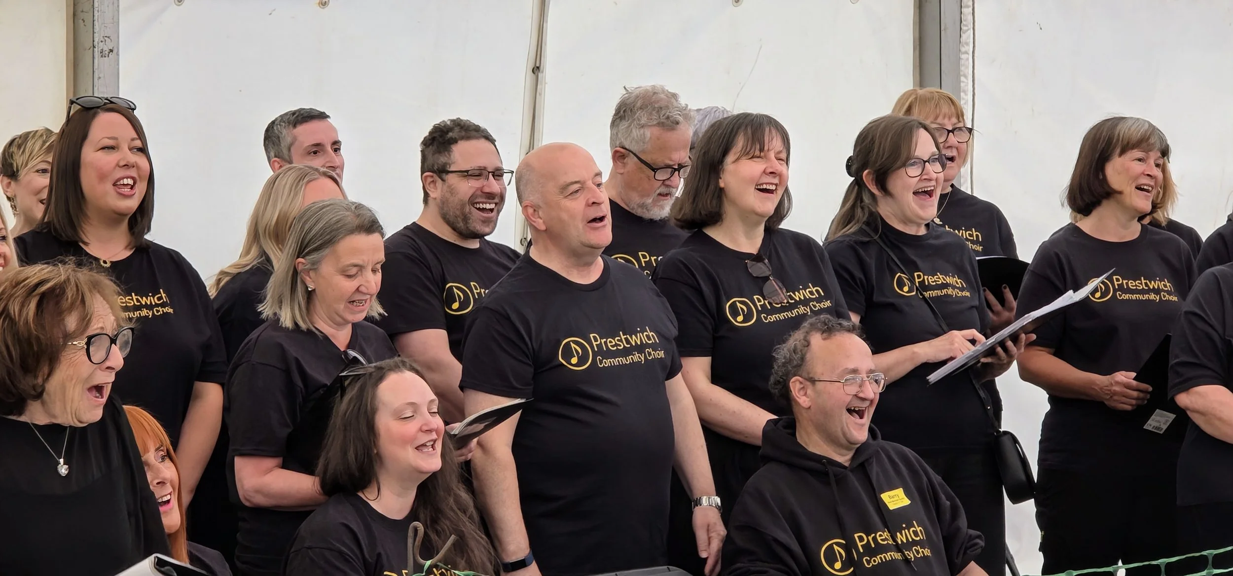 A group of adults wearing black Prestwich Community Choir t-shirts are standing and seated, singing and smiling inside a tent.