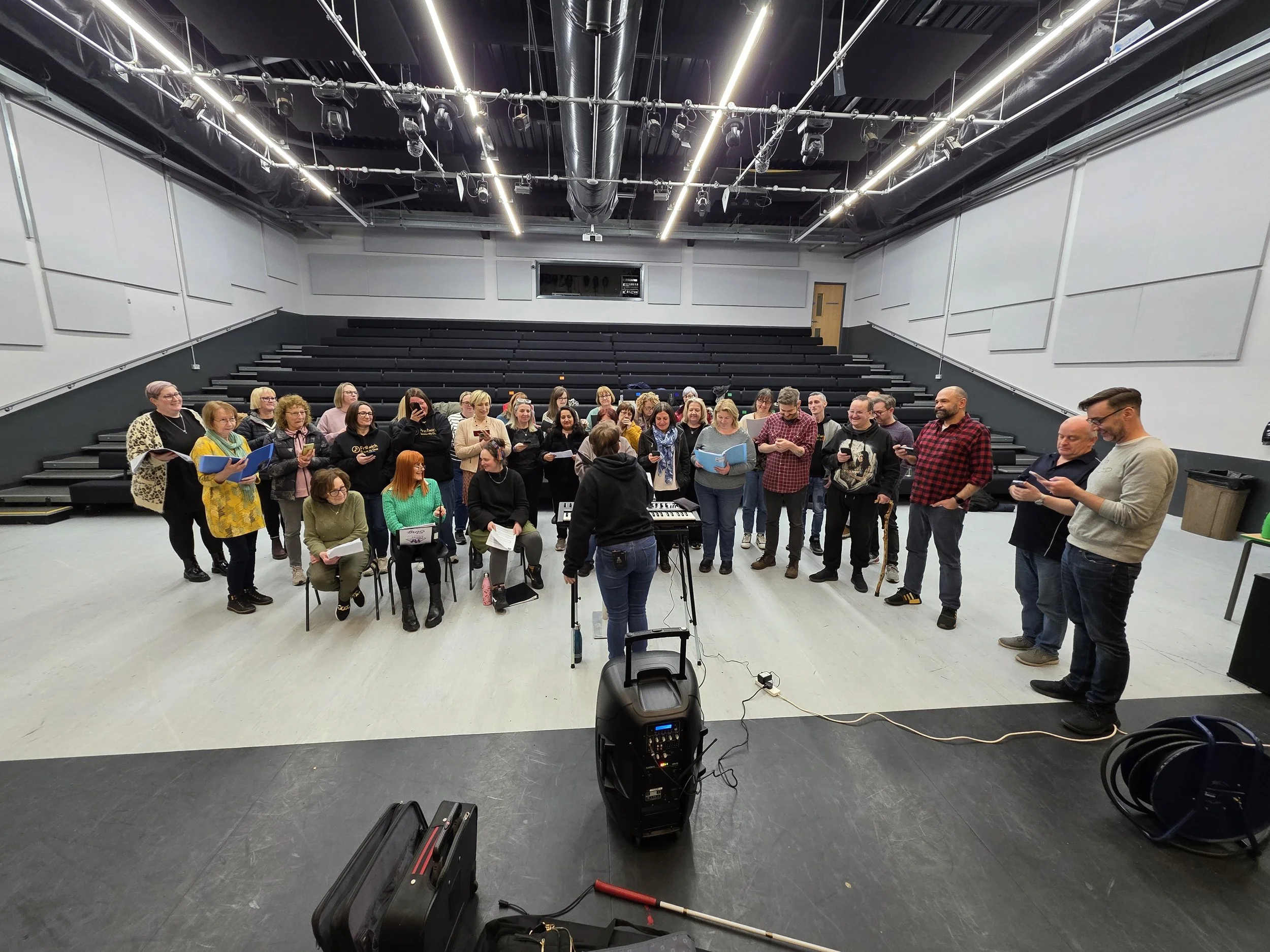 A large group of people gathered in a rehearsal or practice space, possibly singing or rehearsing, with chairs, music sheets, and a keyboard. The space has tiered seating and high ceilings with lights and air ducts.