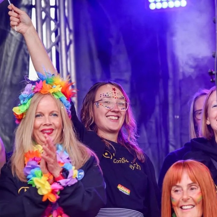 Group of women at a celebration or event, some wearing colorful accessories and face paint, with a stage and lighting in the background.