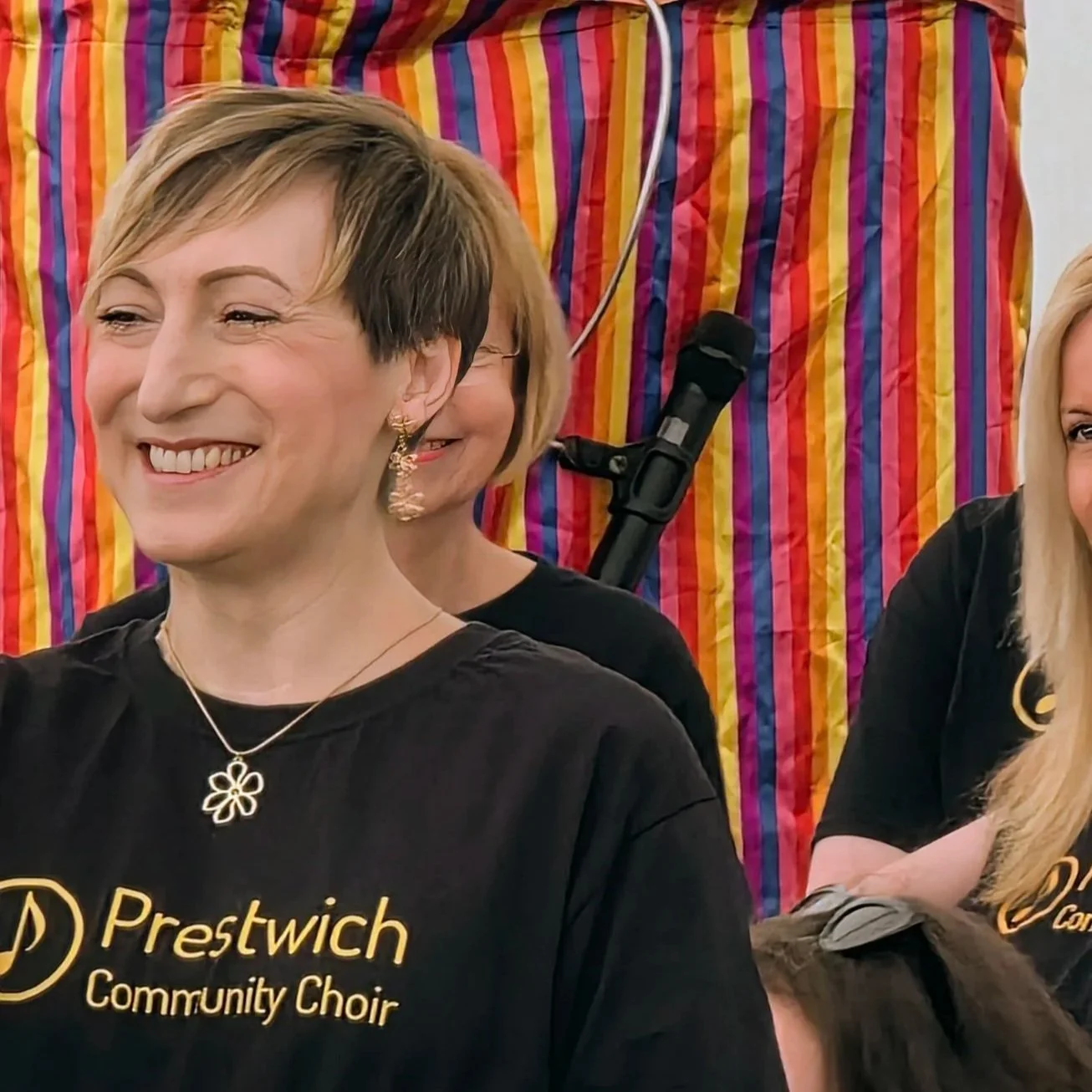 Smiling woman with short hair wearing a black Prestwich Community Choir t-shirt, gold necklace, and earrings, standing in front of a colorful striped backdrop.