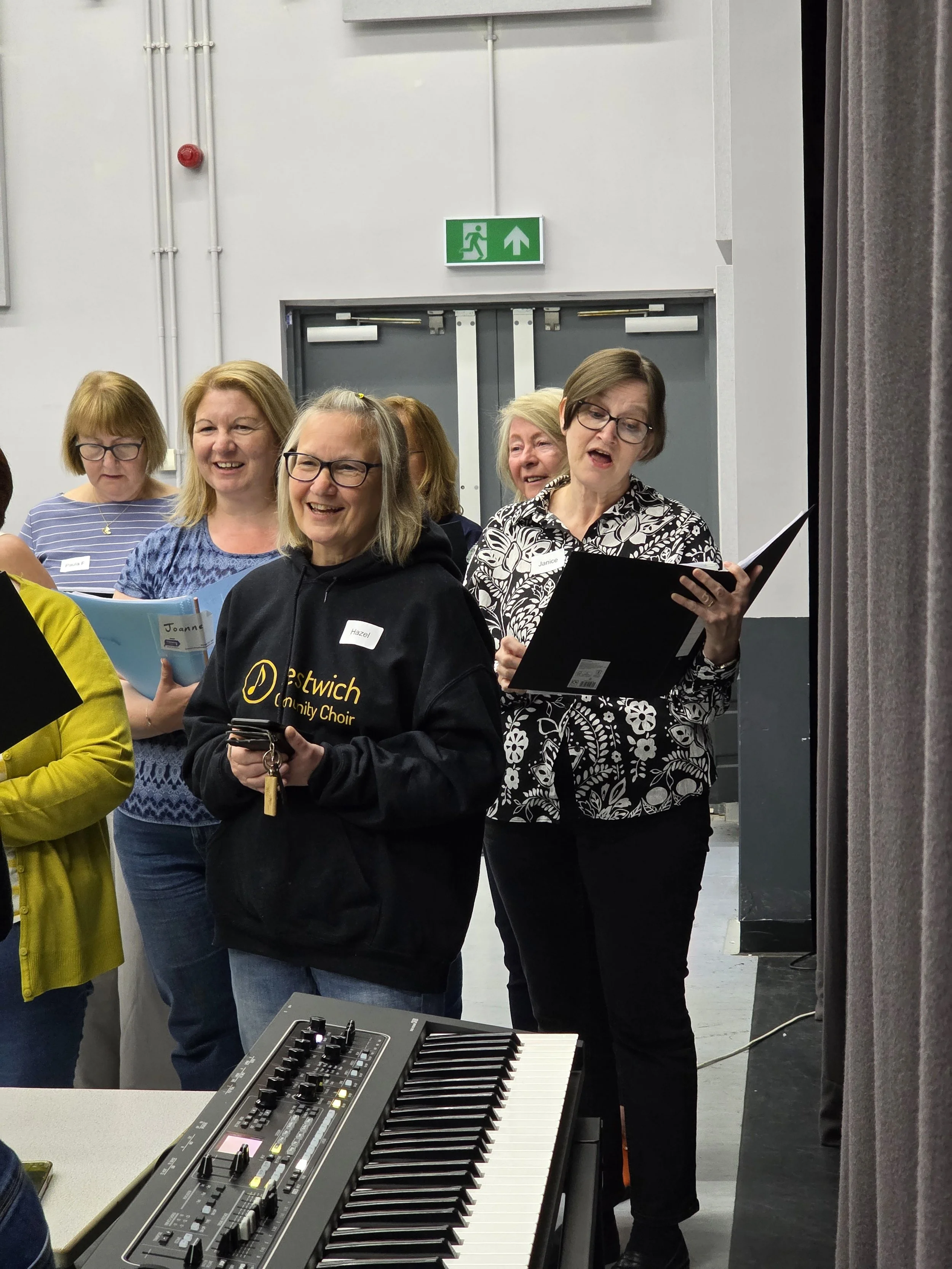 A group of women singing in a choir, holding sheet music, in a room with a keyboard in the foreground, gray walls, and a doorway with an emergency exit sign above it.