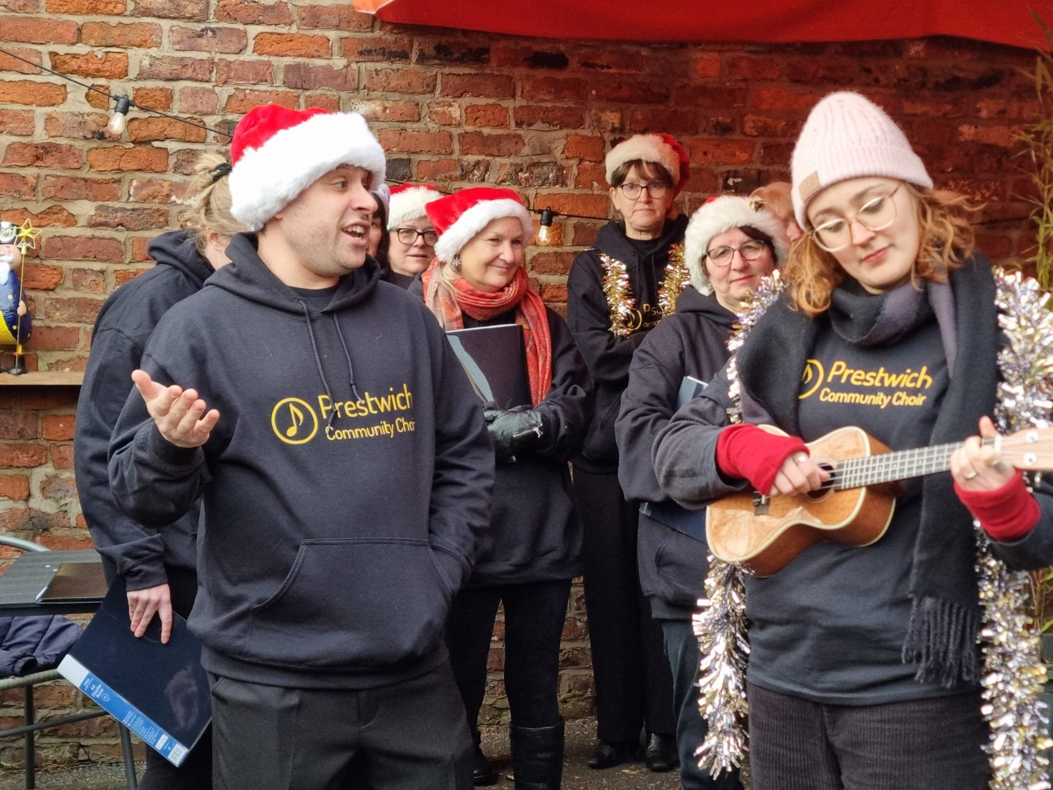 Group of people wearing Christmas hats, some holding songbooks, with a woman playing a guitar, standing against a brick wall decorated for Christmas.