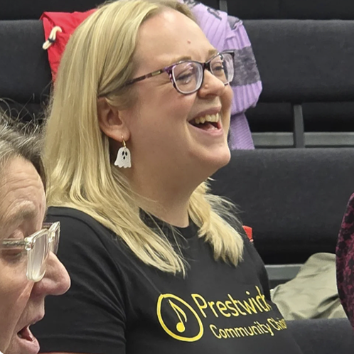 A woman with blonde hair, glasses, and ghost-shaped earrings smiling and laughing in a group setting.