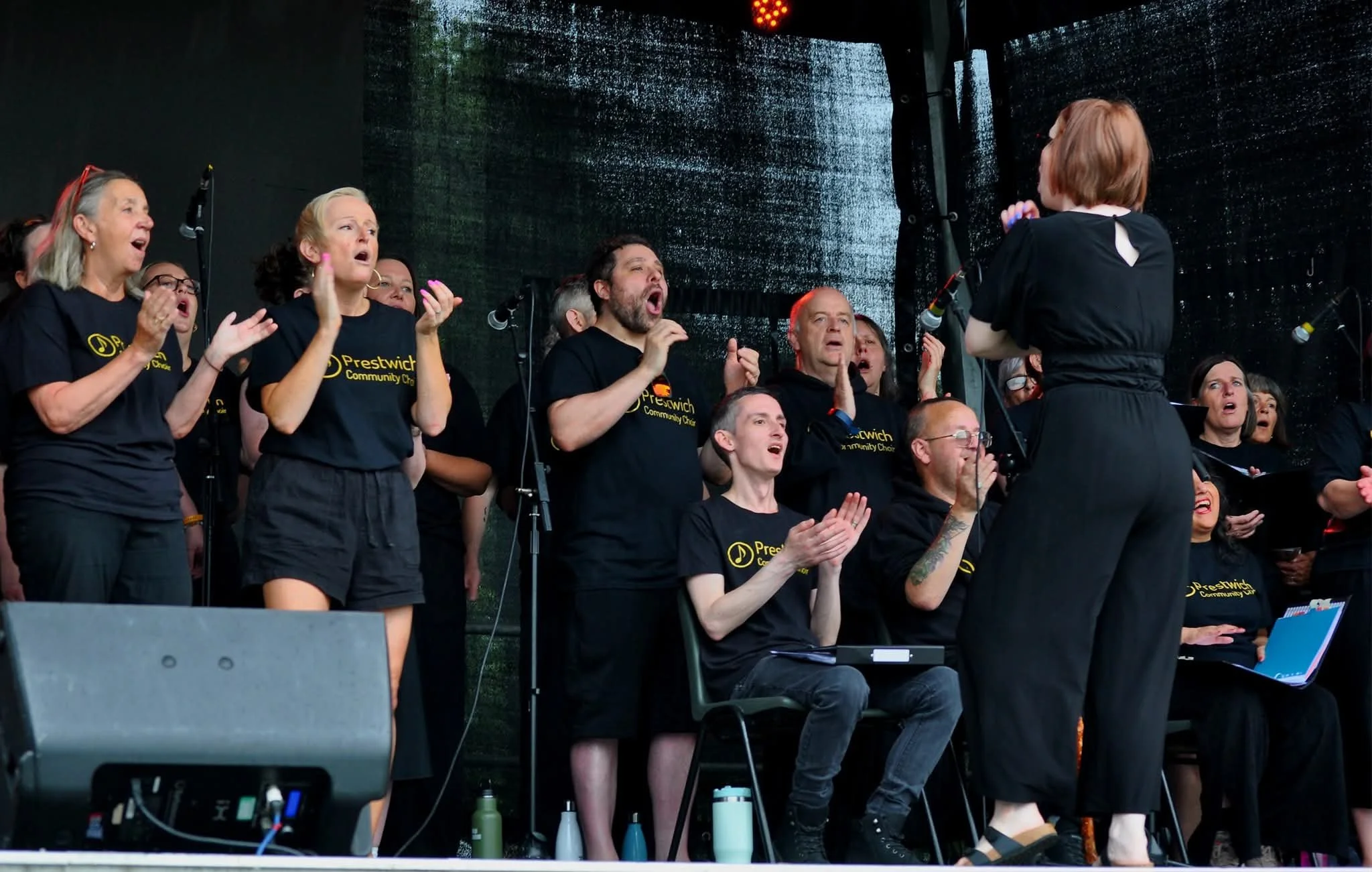 A choir of diverse men and women singing on stage during a performance, led by a conductor with their back to the camera, in an outdoor concert setting.