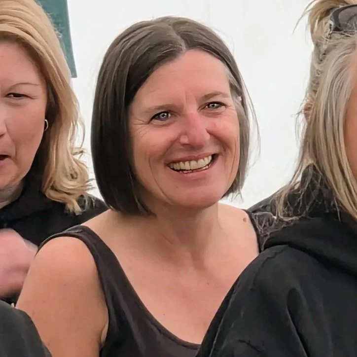 Smiling woman with short dark hair and a sleeveless black top, standing among other women.