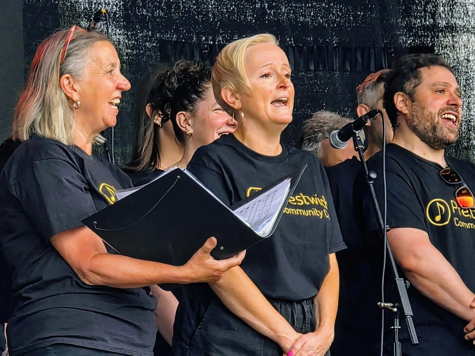 Group of people singing or speaking on stage, wearing black T-shirts with yellow text, some holding papers, smiling and standing near a microphone.