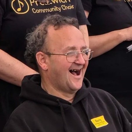 A man with glasses and curly hair laughing, wearing a black hoodie and a yellow name tag, in a group setting.