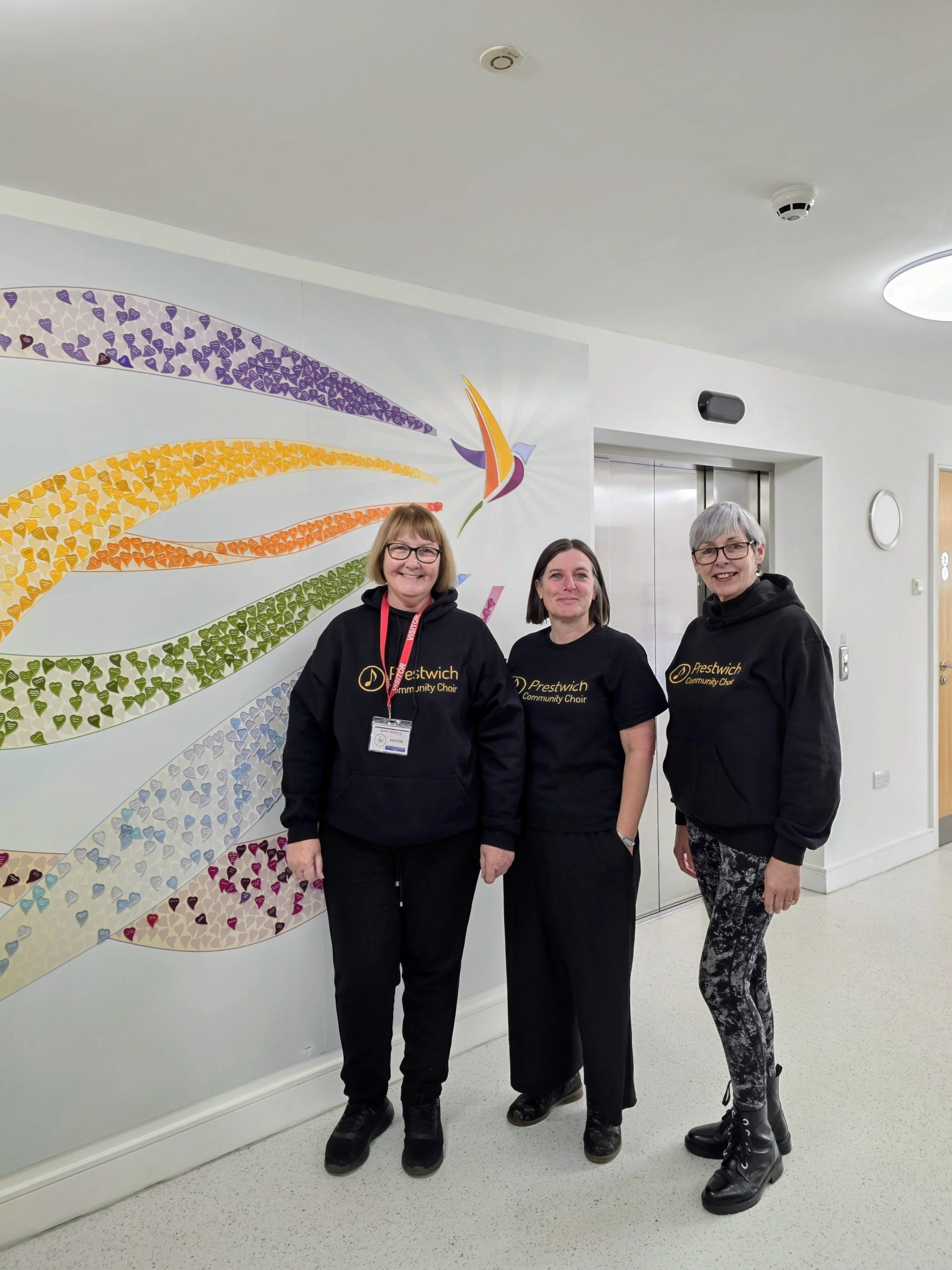 Three women standing in front of a colorful wall art mural, wearing black hoodies with 'Prestwich Community Choir' printed on them, smiling at the camera.