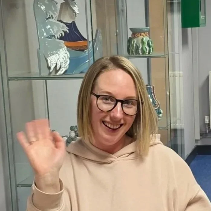 Smiling woman with glasses waving, in an indoor space with display shelves and decorative items in the background.