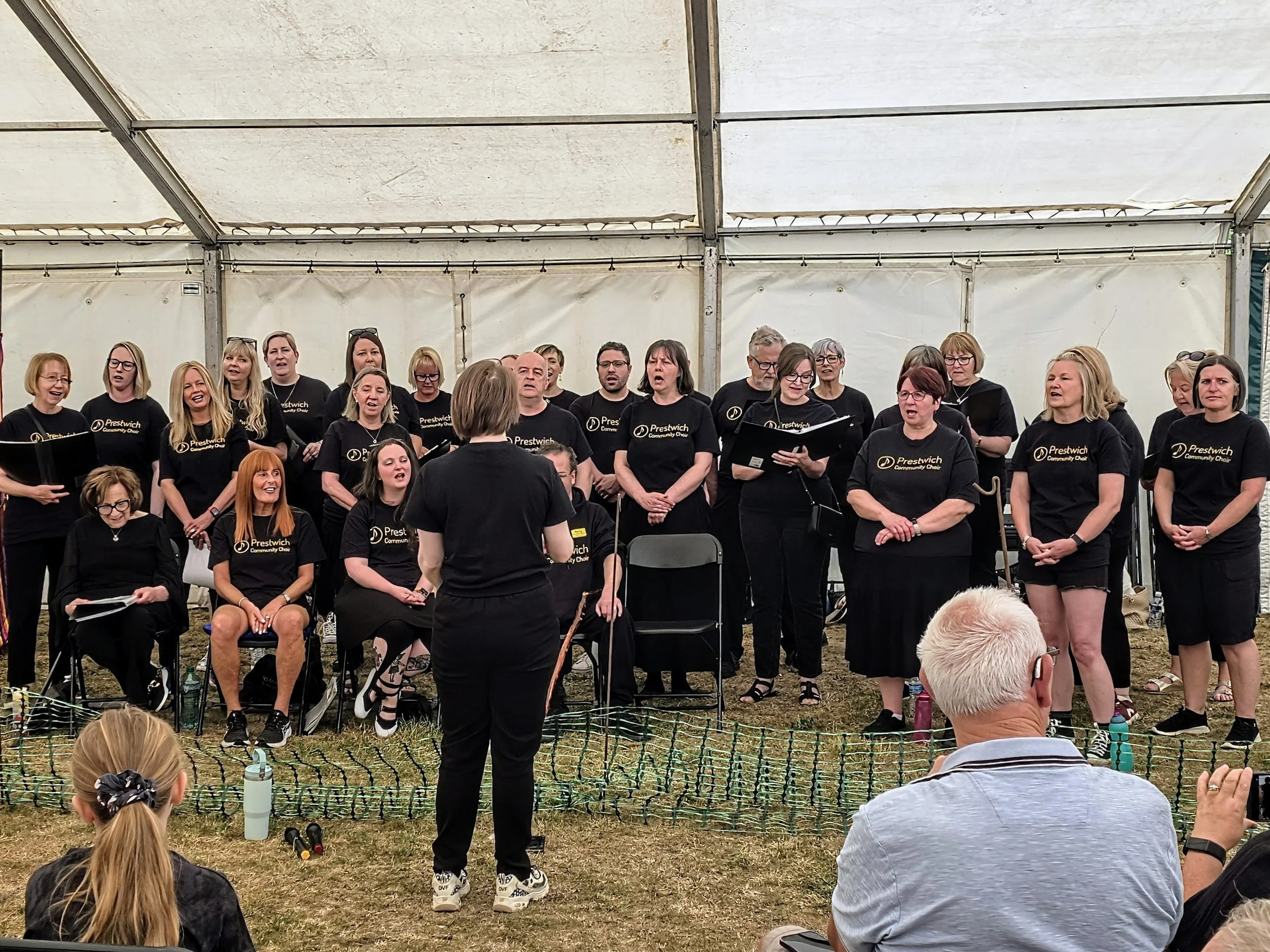 A choir of adults performing under a white tent, led by a conductor, with some members wearing black shirts that read 'Prestwich Community Choir.'