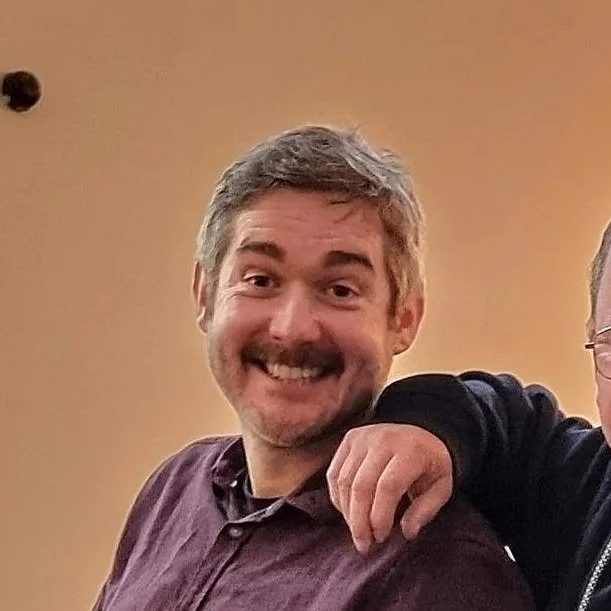 A smiling man with gray hair and facial hair, wearing a maroon shirt, is in an indoor setting with a beige wall background. Part of another person is visible to his right.