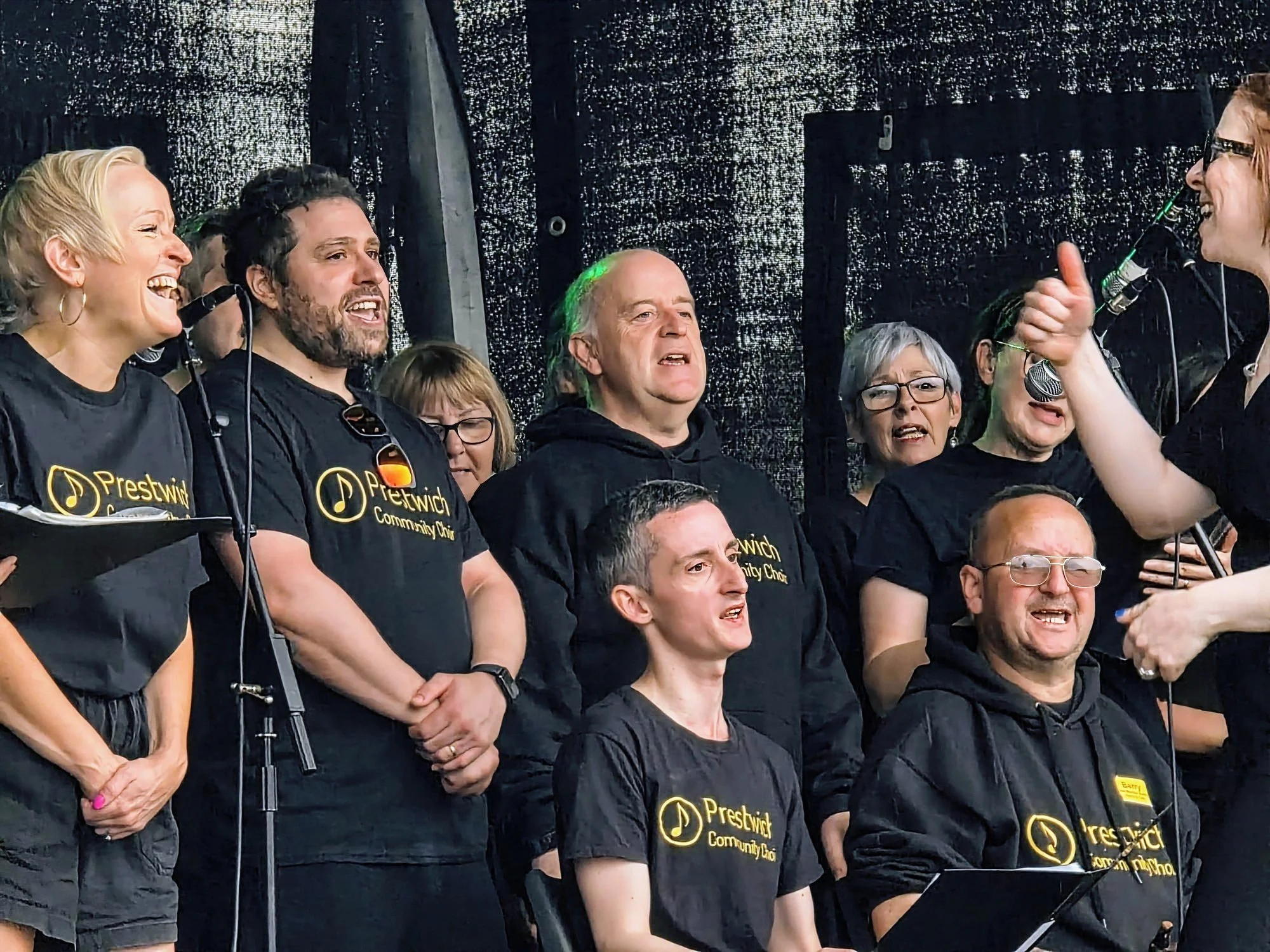Group of people singing on stage, wearing black Preswich Community Choir shirts.