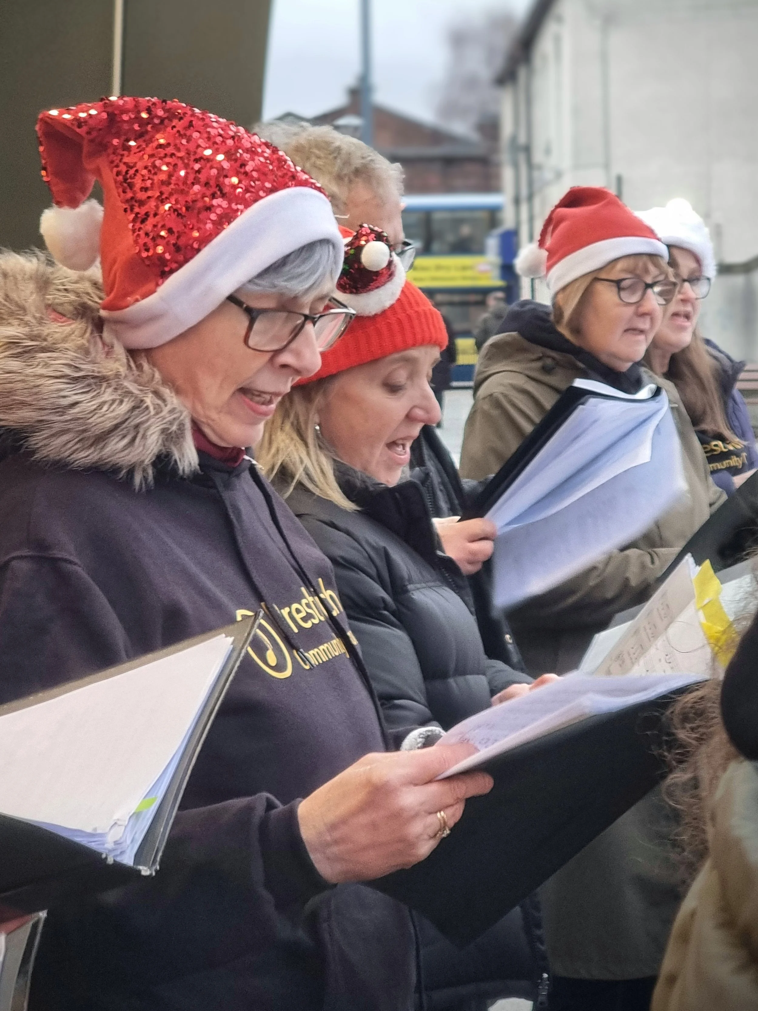 Group of women singing Christmas songs while holding songbooks, wearing festive Santa hats and winter coats outdoors.