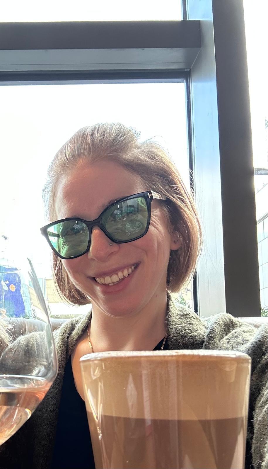 A woman with short blonde hair and sunglasses smiling at a camera, with a glass of white wine and a latte in front of her, sitting by a window.