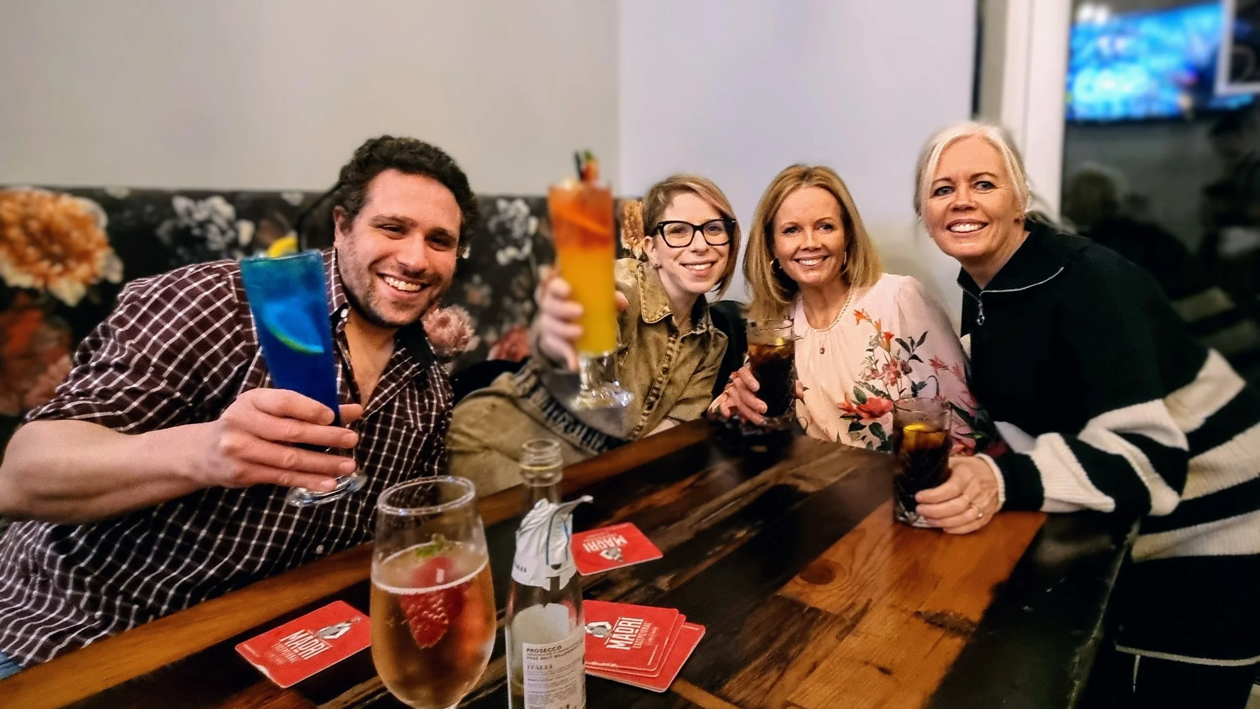 Five people sitting at a bar holding colorful drinks, smiling, with a wooden table and a floral patterned wall in the background.