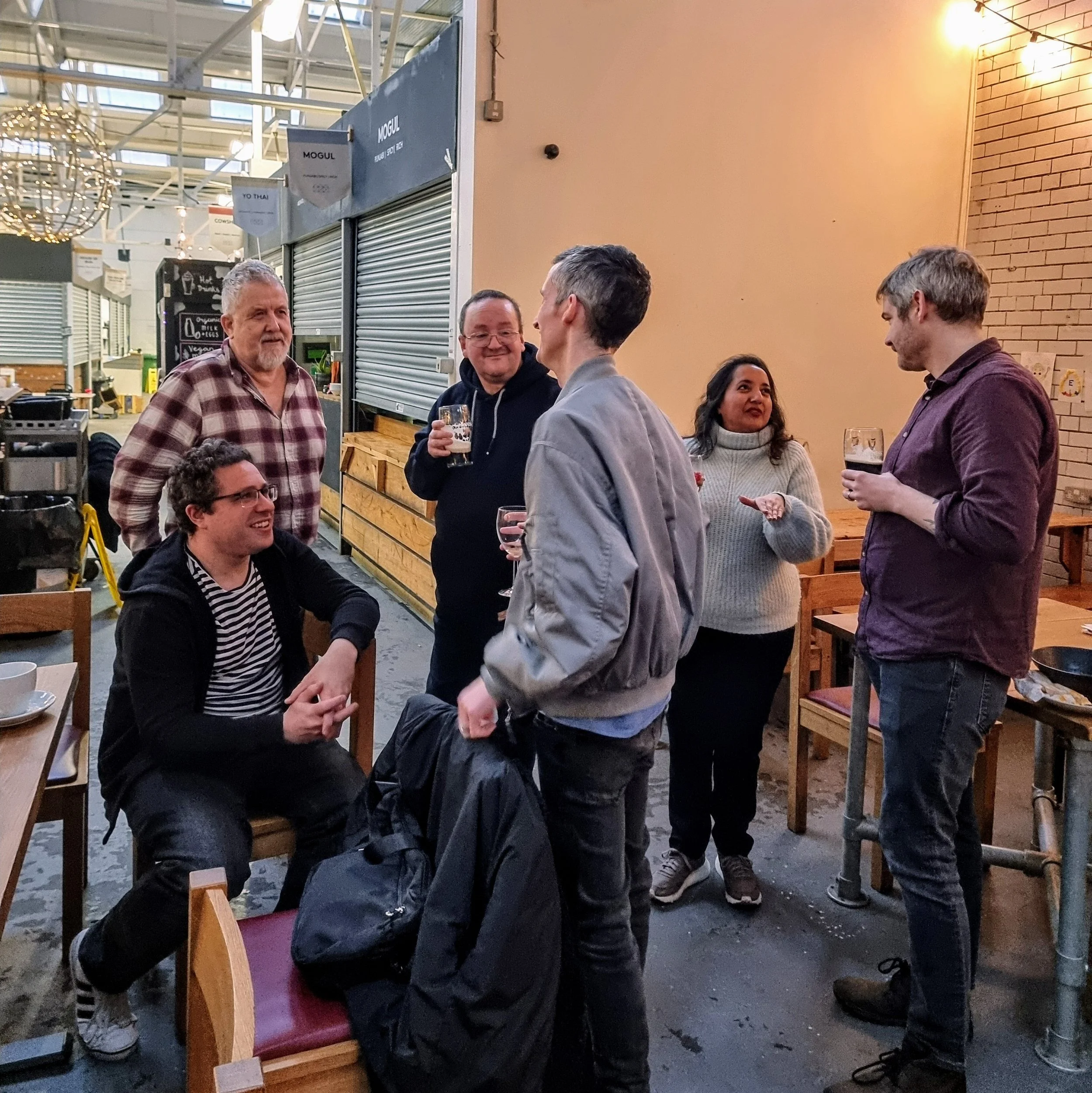Group of six people socializing at a casual indoor setting, four standing and two seated, with drinks in hand, smiling and engaging in conversation.