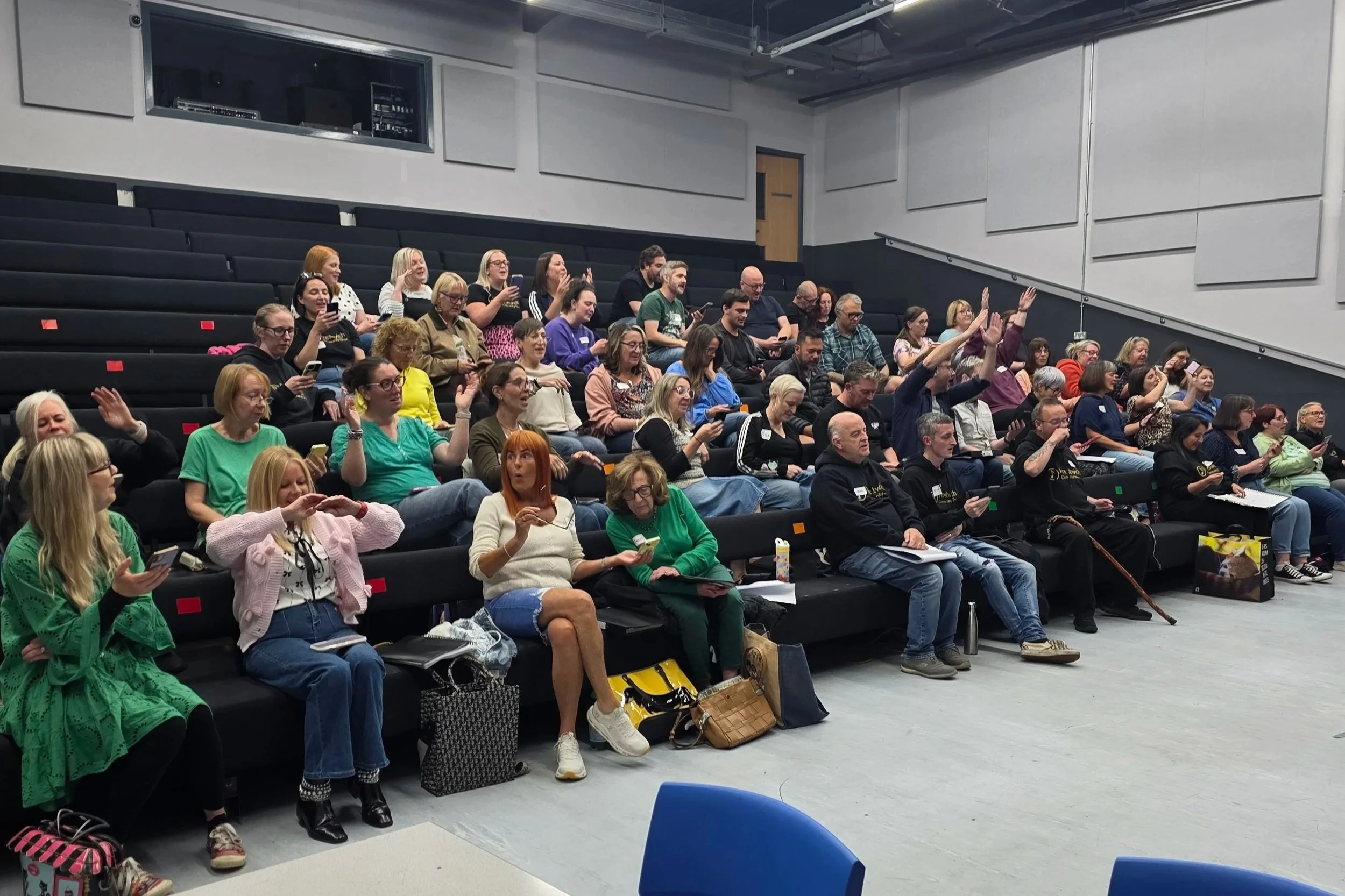 Audience in a lecture hall participating in a voting or question-answer session, with some raising their hands and others using mobile devices.