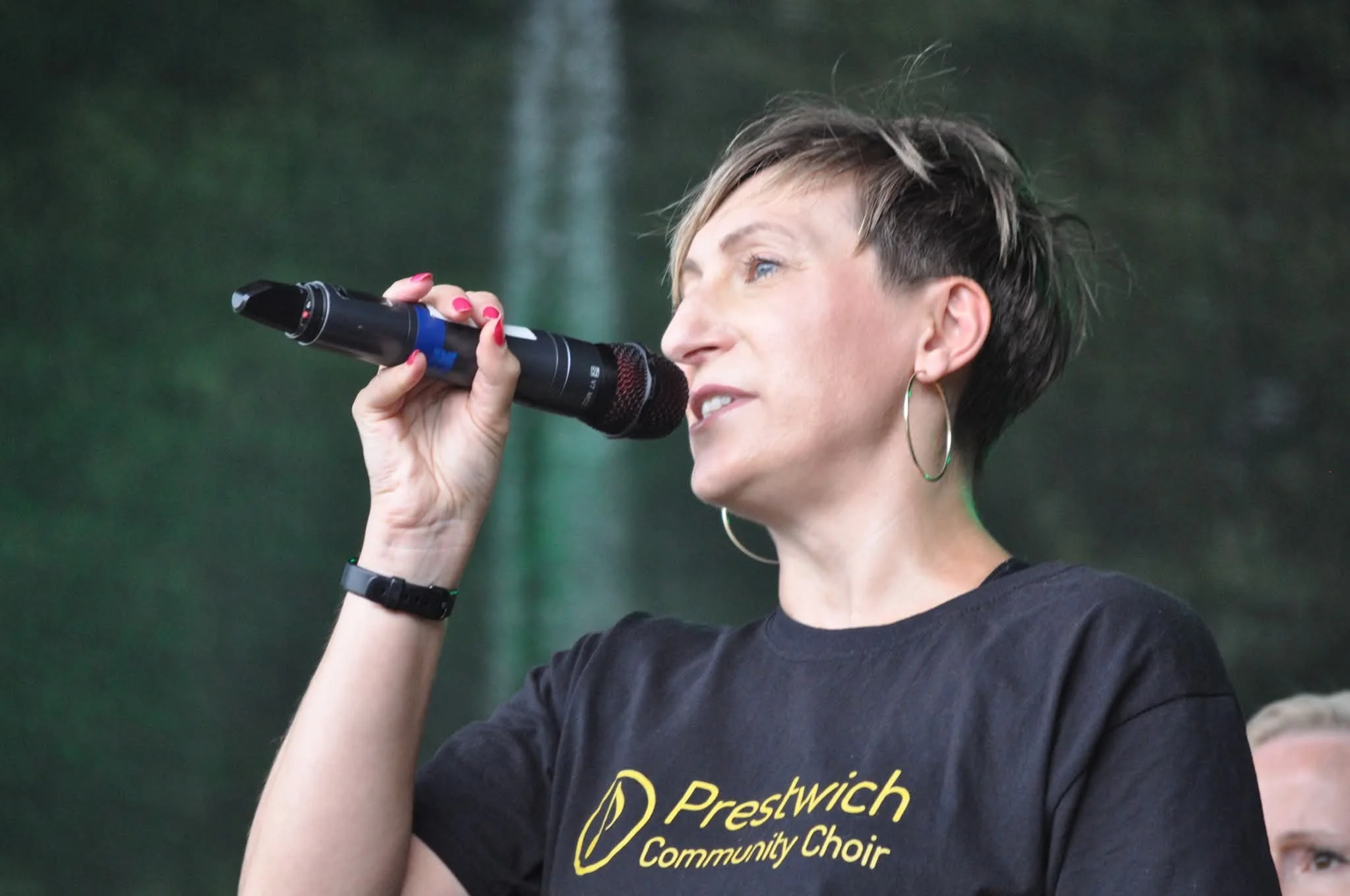 A woman with short hair, wearing a black t-shirt that says 'Prestwich Community Choir,' is holding a microphone and singing in front of a greenish background.