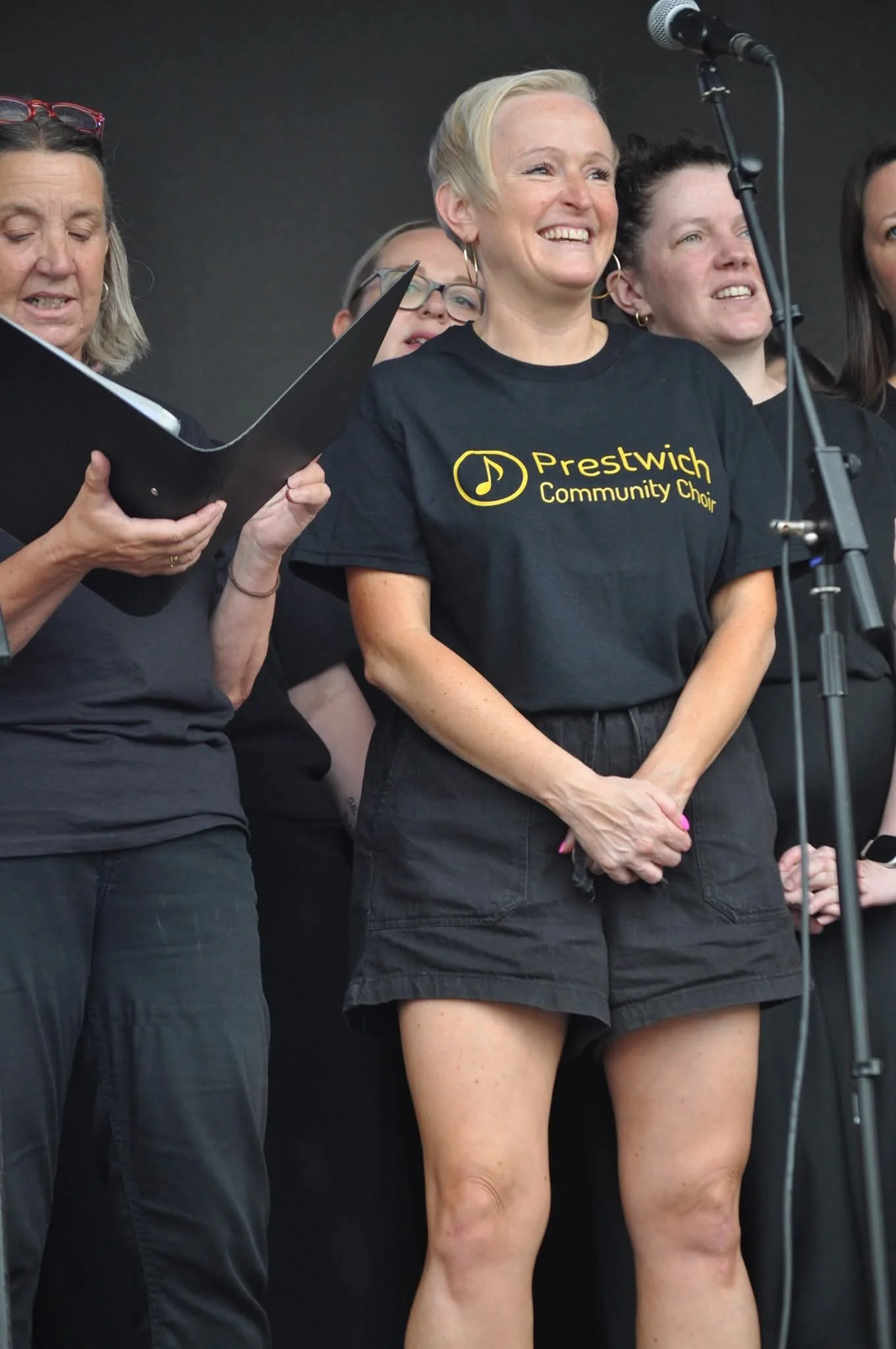 Group of women on stage, one wearing a black T-shirt with yellow text and logo for Prestwich Community Choir, standing behind a microphone, smiling, with others around her, holding a music folder.