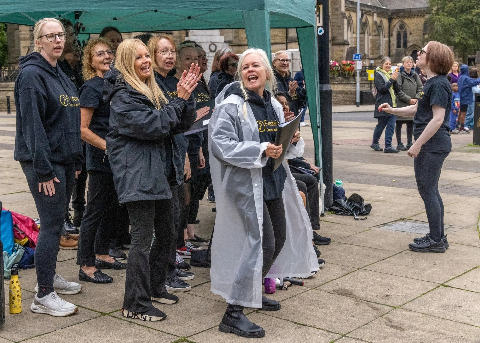 A group of women singing in a choir outdoors, with one woman leading and holding sheet music, under a green canopy, in front of a historic stone building, on a cloudy day.