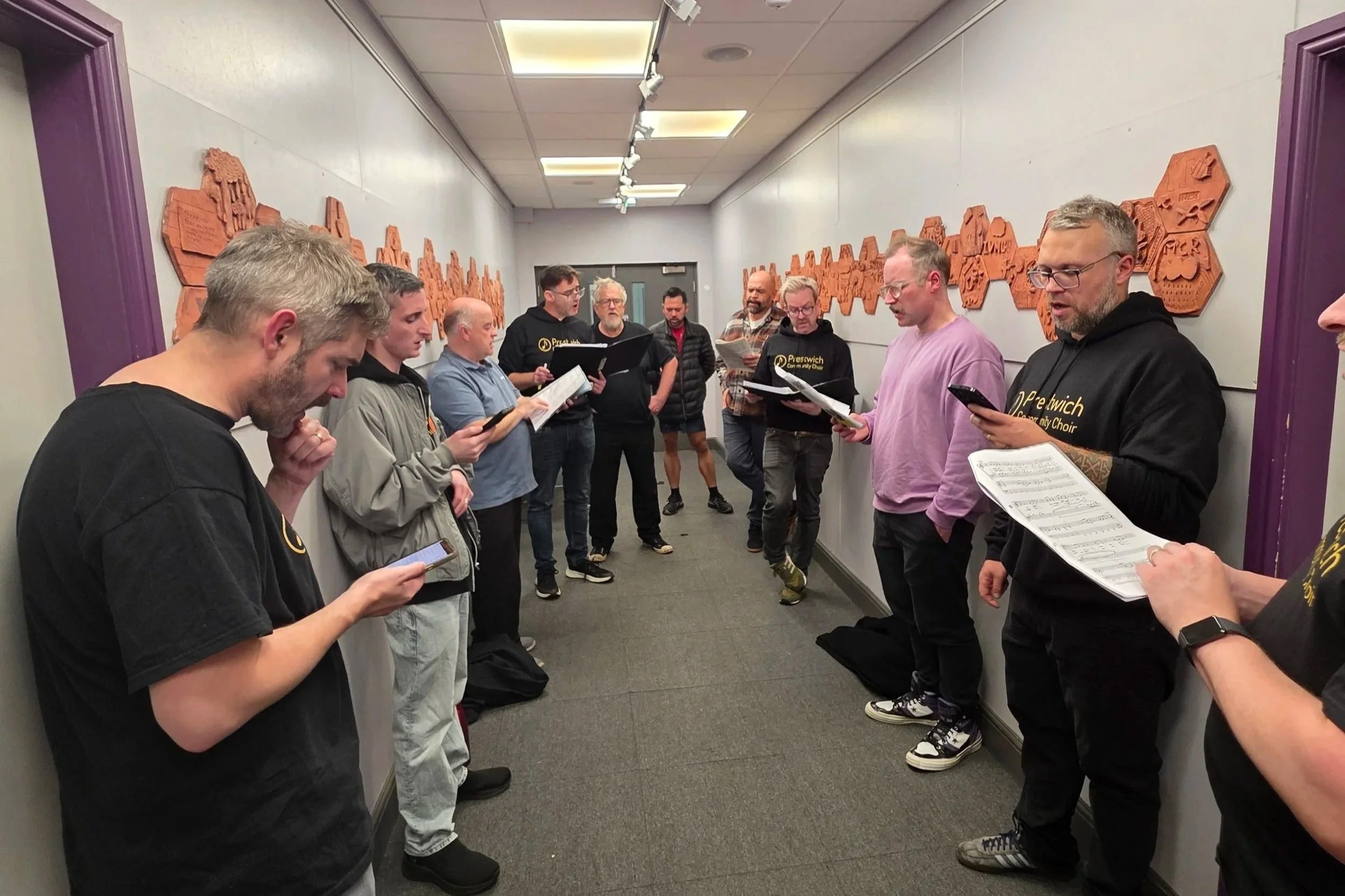 Group of people standing in a hallway, singing from sheet music and using mobile devices, with orange artwork on the walls.