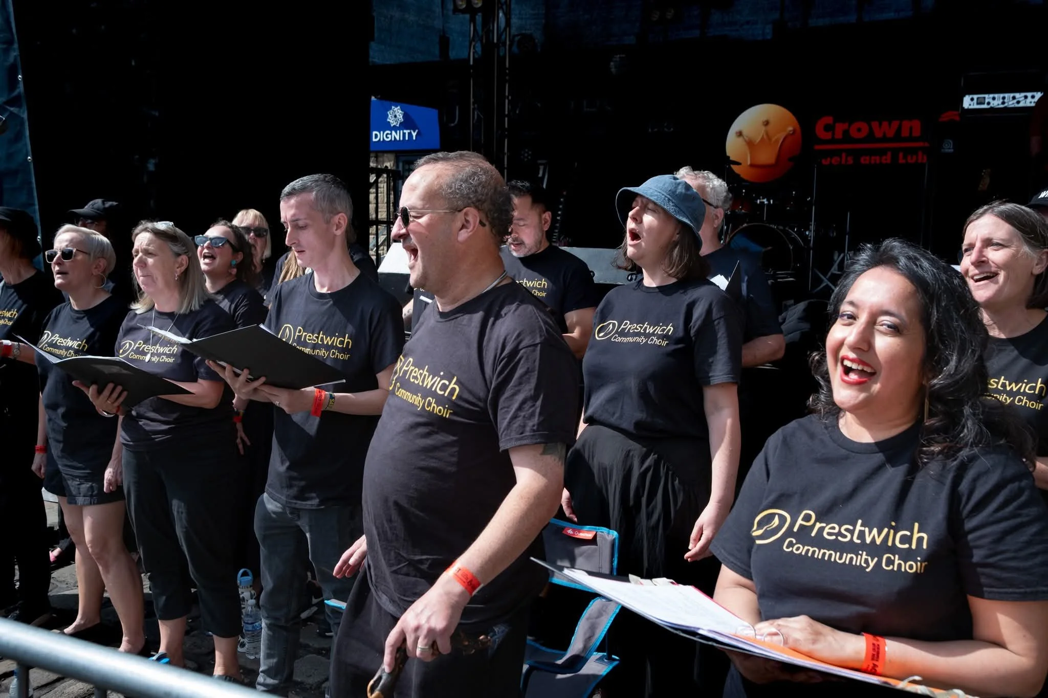 Group of people singing in a choir outdoors, wearing black shirts with yellow text that reads 'Prestwich Community Choir', some holding songbooks, with a stage and banners in the background.