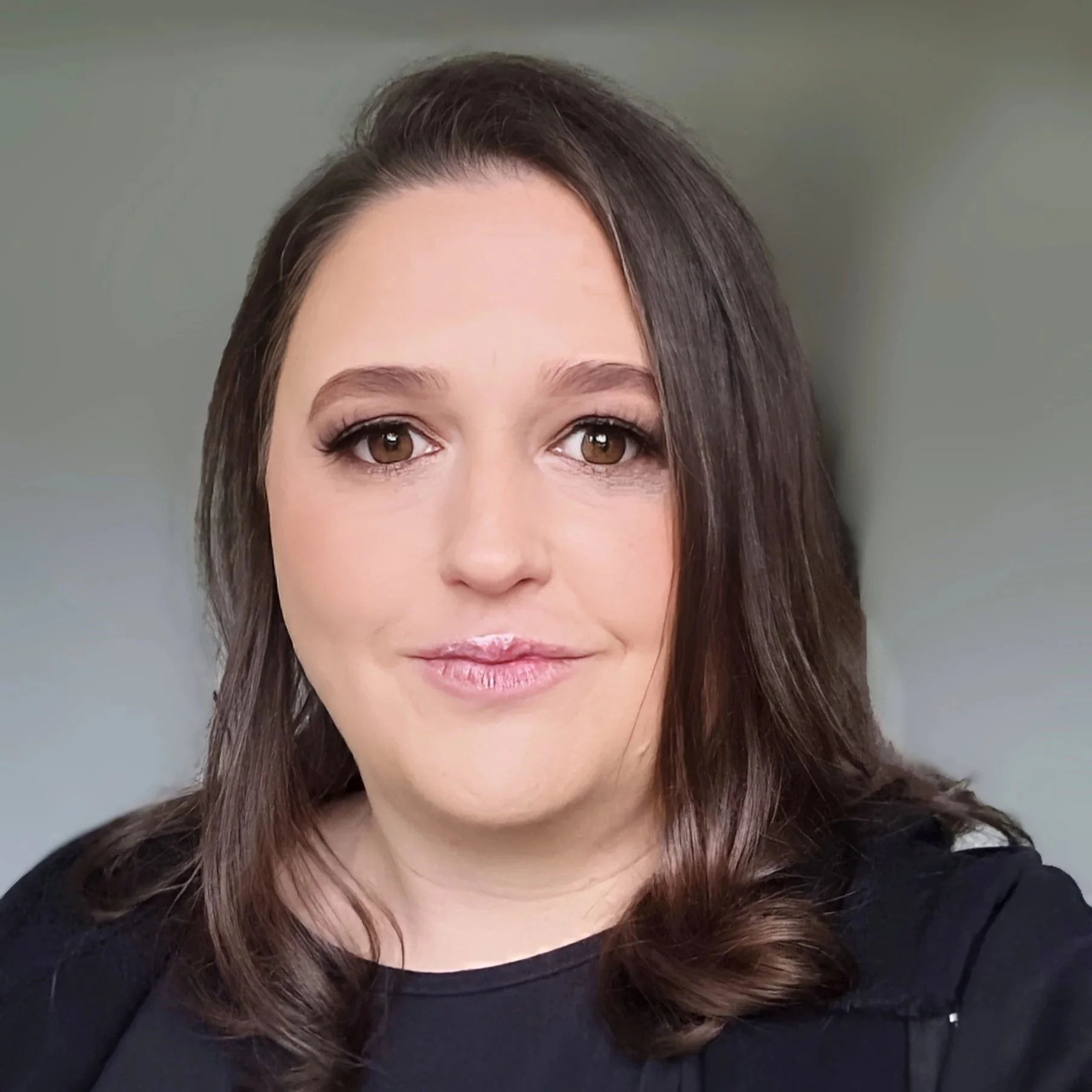 Portrait of a woman with brown eyes, long brown hair, wearing makeup and a black top, smiling softly.