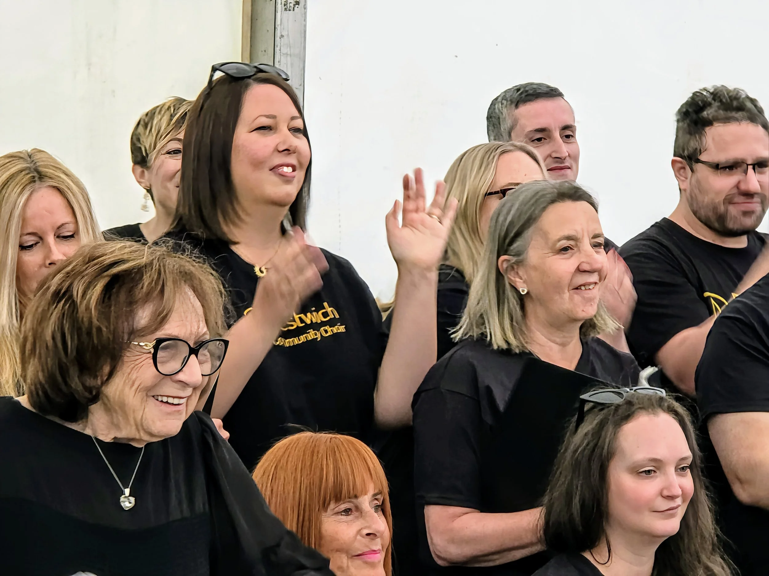 Group of people at an indoor event, some smiling and others raising hands to clap, wearing black shirts, in front of a white wall.