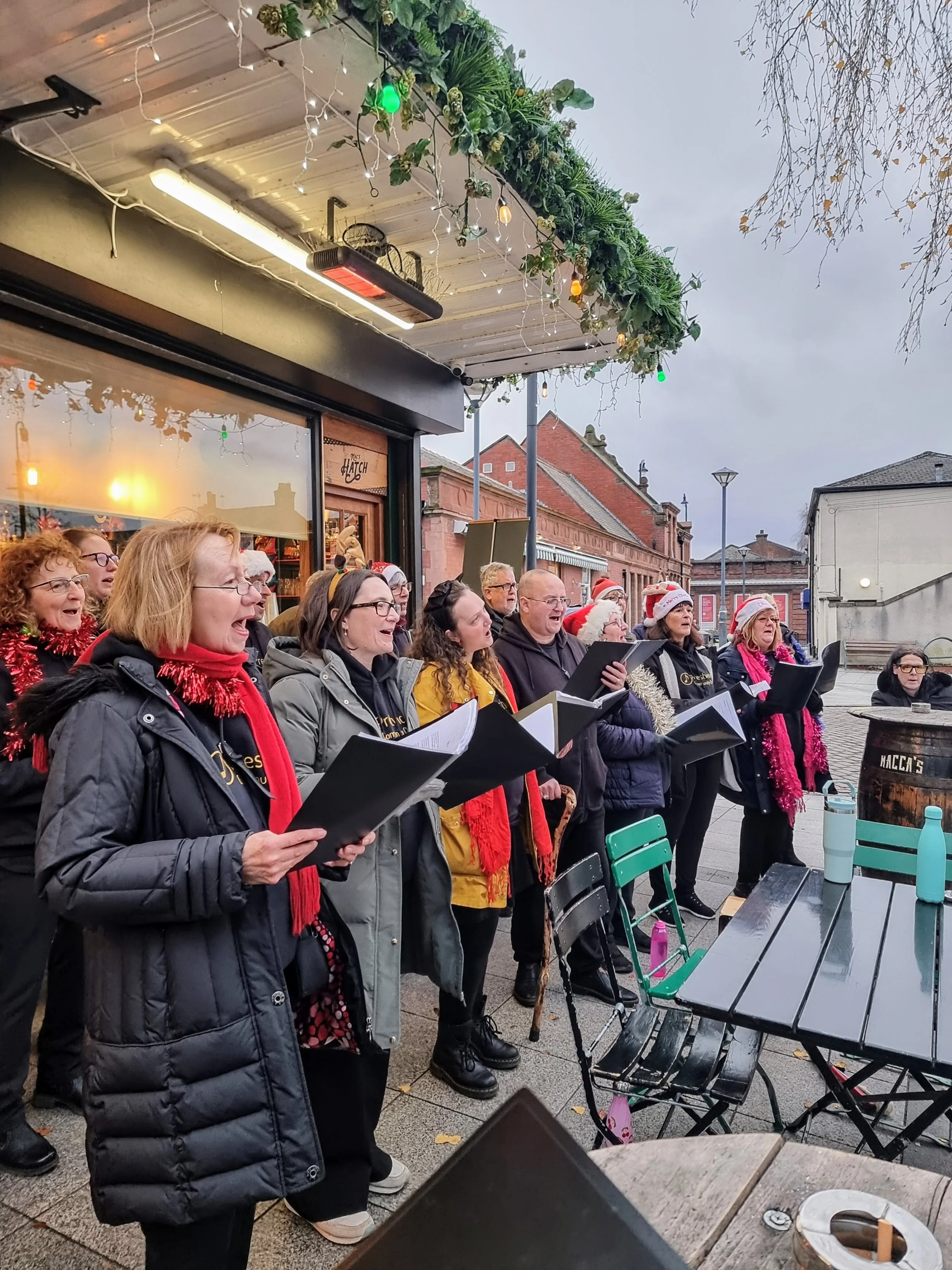 A group of people singing Christmas carols outdoors in front of a shop decorated with holiday lights and greenery, some wearing Santa hats and holiday scarves.