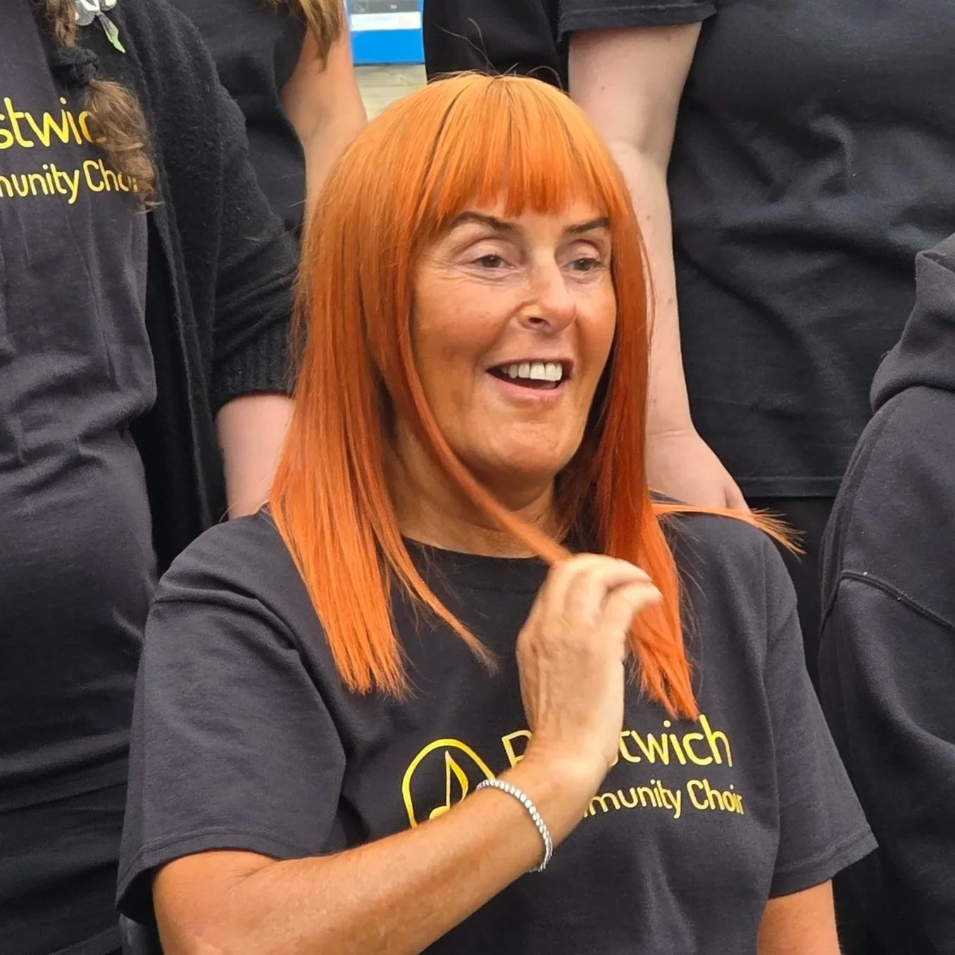 A smiling woman with bright orange hair wearing a black shirt with yellow text, surrounded by other people wearing similar black shirts.