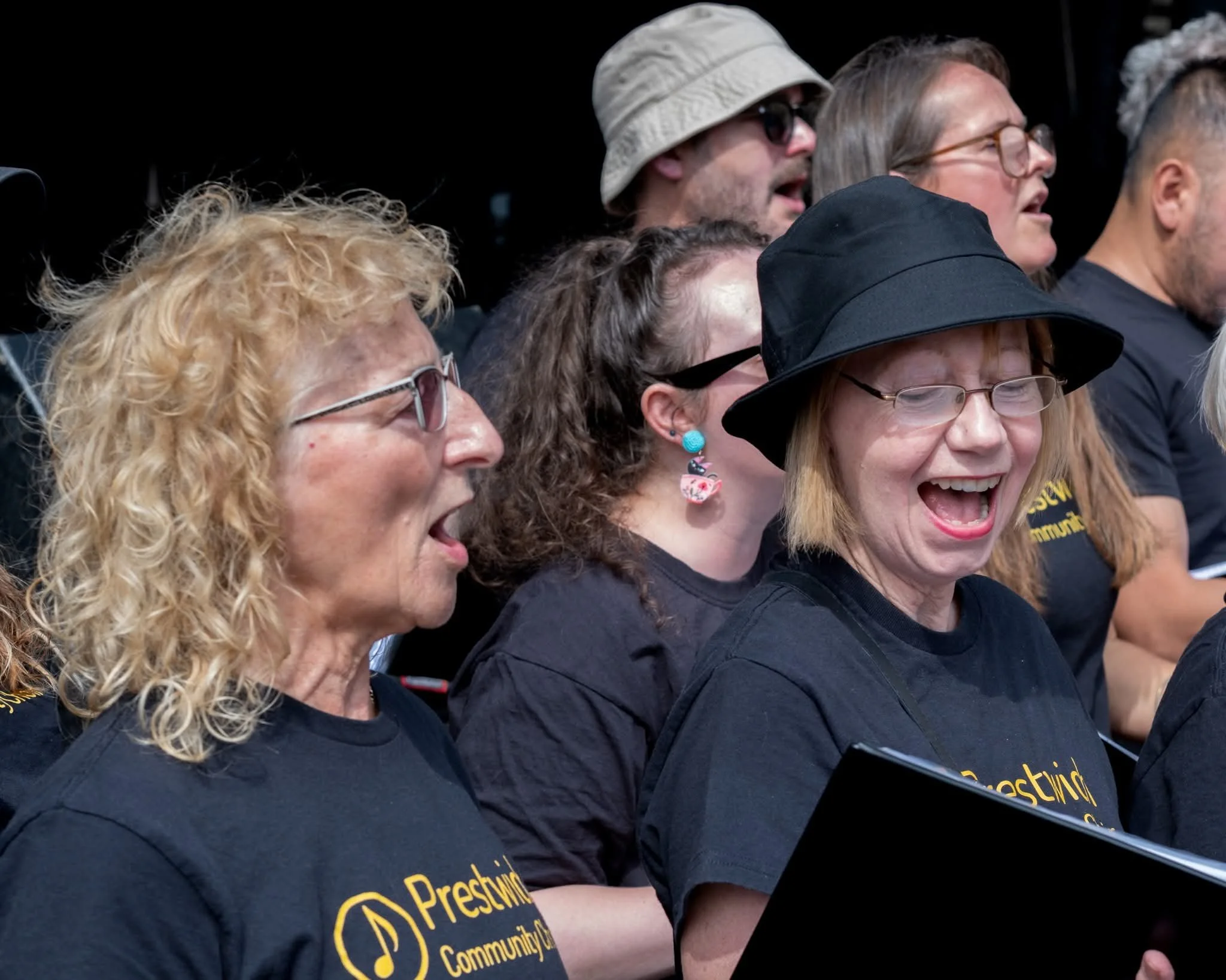 Group of people singing or cheering, wearing black shirts with yellow text, some with glasses and hats, displaying expressions of joy and enthusiasm.