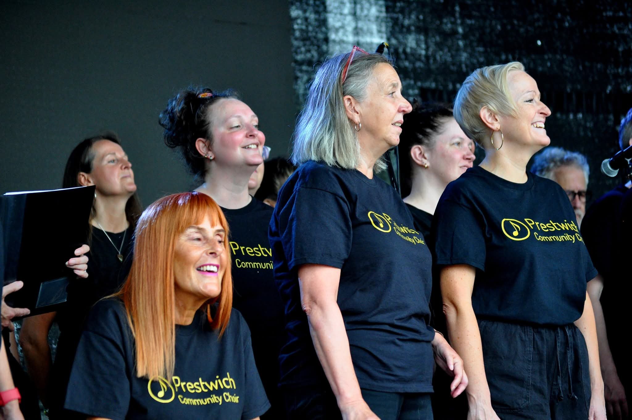 A group of women singing in a choir, wearing black t-shirts with a yellow logo and text that reads 'Preston Community Choir'.