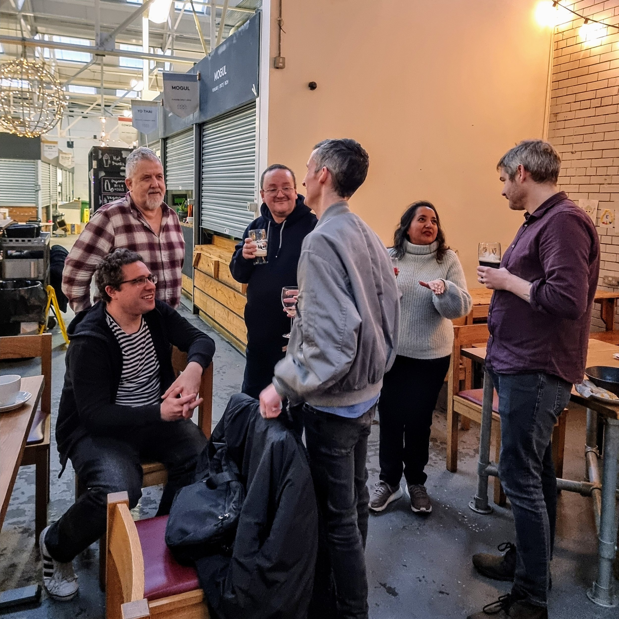 Six people standing and sitting in a casual indoor dining area, engaging in conversation and holding drinks.