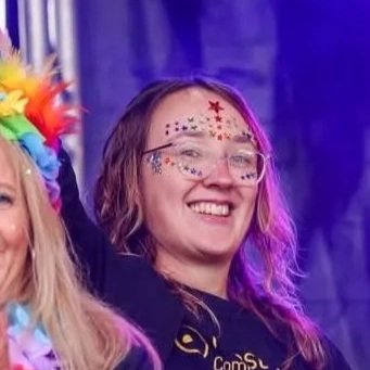 A woman wearing glasses, smiling, decorated with colorful star and dot face paint, and a rainbow feathered headband, at a festive event.