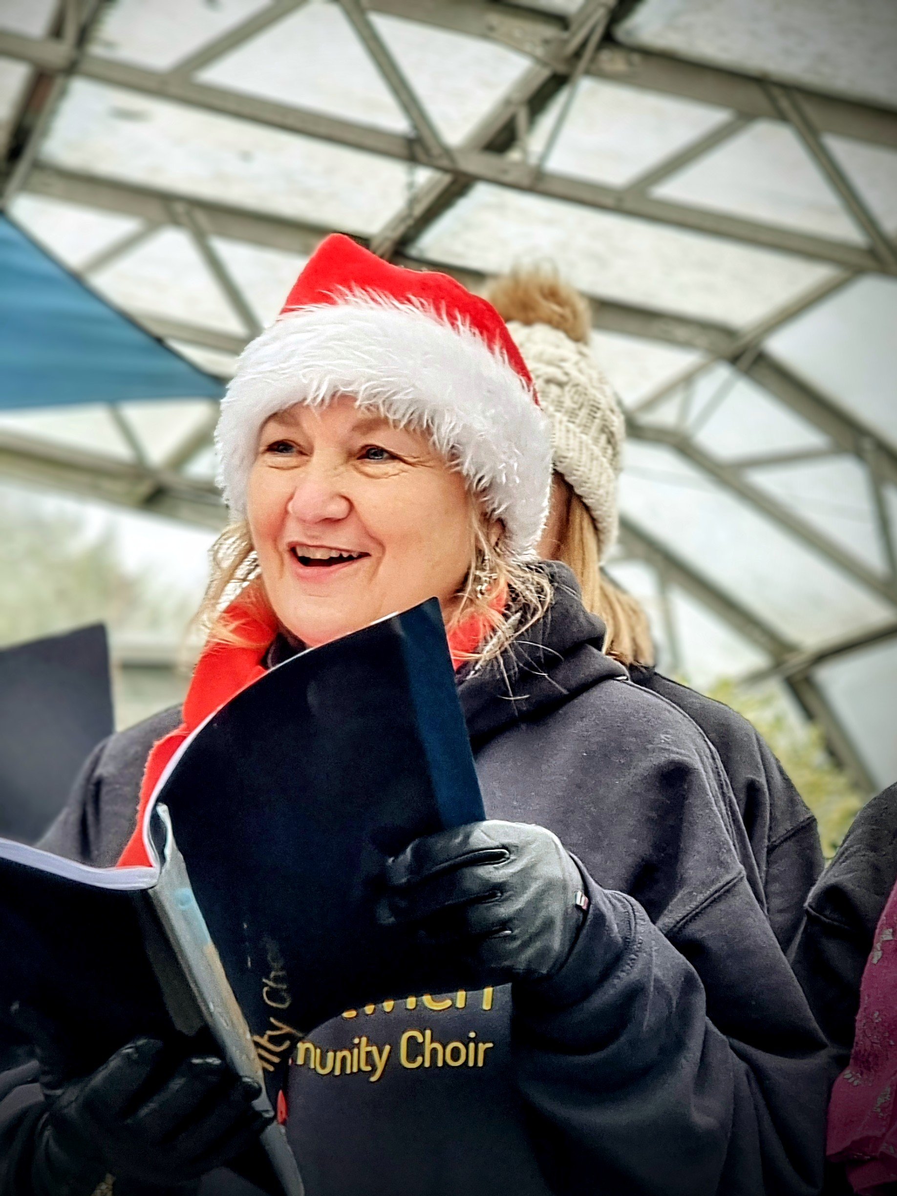 Woman wearing a Santa hat and gloves, smiling and holding a booklet, outdoors under a metal structure.