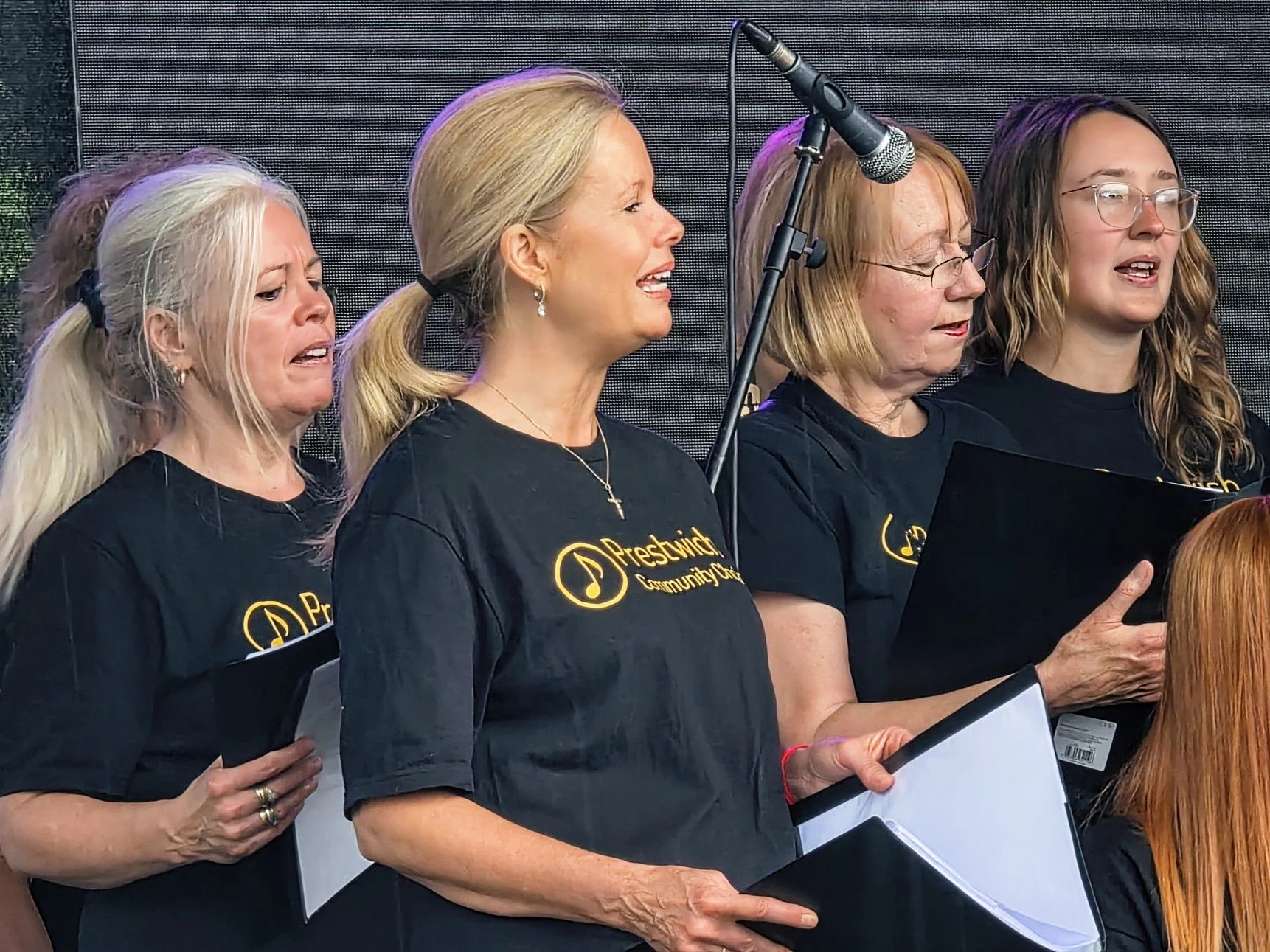 Group of four women singing or performing, holding music sheets, wearing black T-shirts with yellow text, and standing in front of a microphone on a stage.