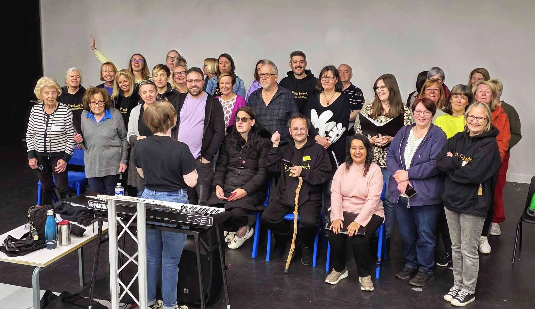 Group of diverse people gathered in a room, some sitting and some standing, smiling and posing for a photo with musical equipment and water bottles on a table in the foreground.