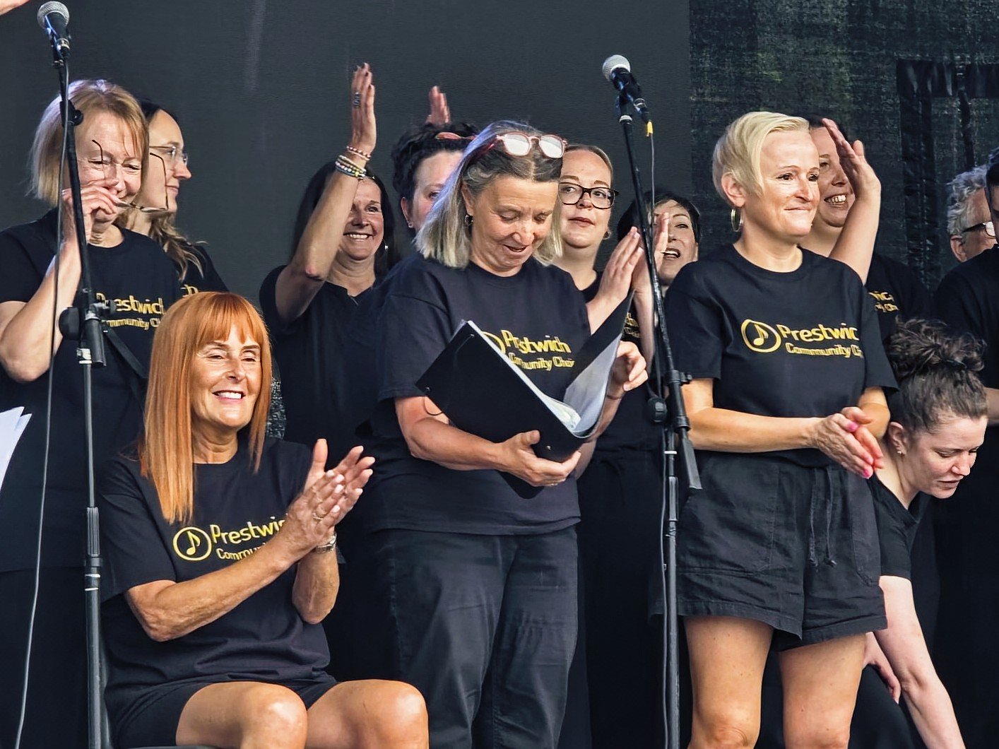 Group of women at a stage, wearing black shirts with yellow text, one seated and clapping, others standing, some smiling, some clapping, in a celebratory and supportive atmosphere.