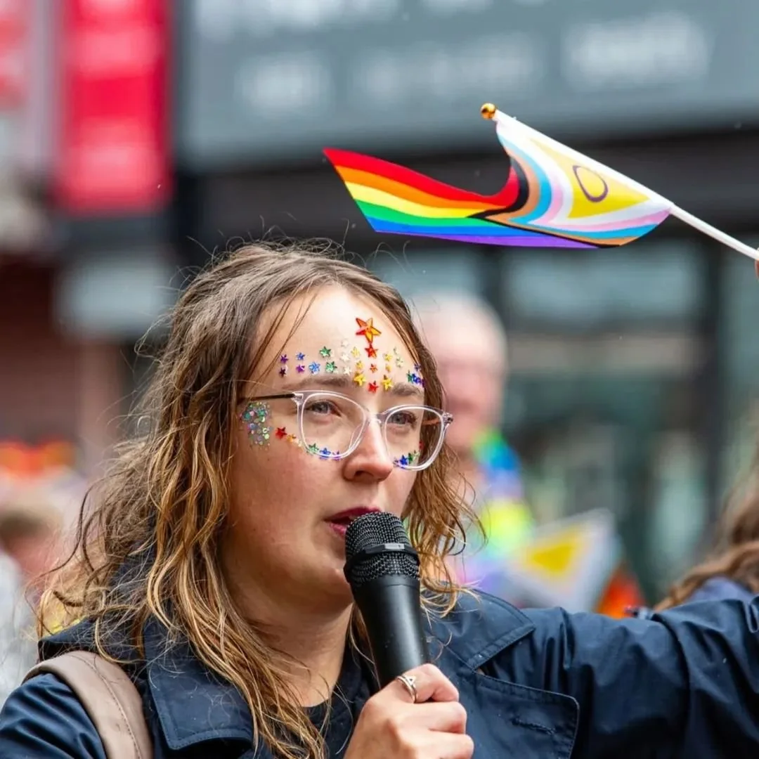 A woman with rainbow star and colorful sticker face decorations, wearing glasses, holding a microphone, and celebrating at a pride event with rainbow pride flags in the background.