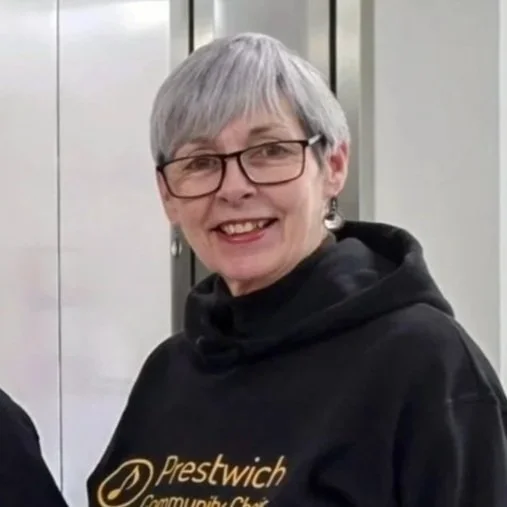 A woman with short gray hair and glasses, smiling and wearing a black sweatshirt with gold writing, standing indoors near a mirrored elevator door.