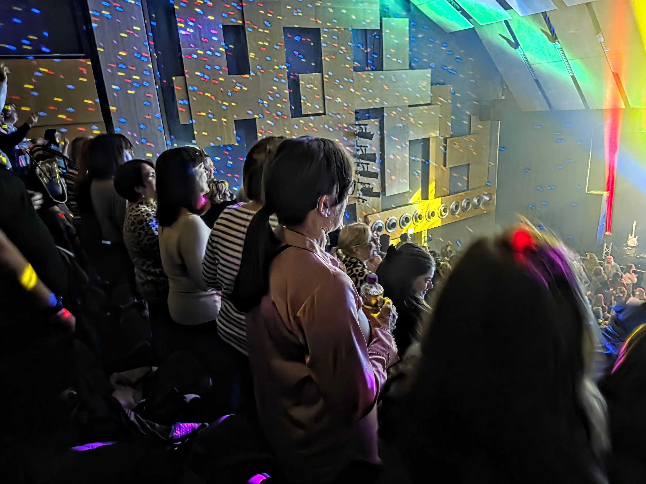 People sitting in a dark concert hall watching a stage illuminated with colorful lights and speakers, with a large wall displaying a visual display.