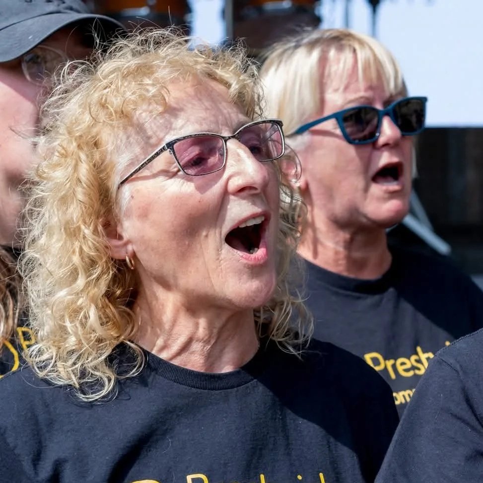Three older women with glasses singing or shouting during a gathering or event.