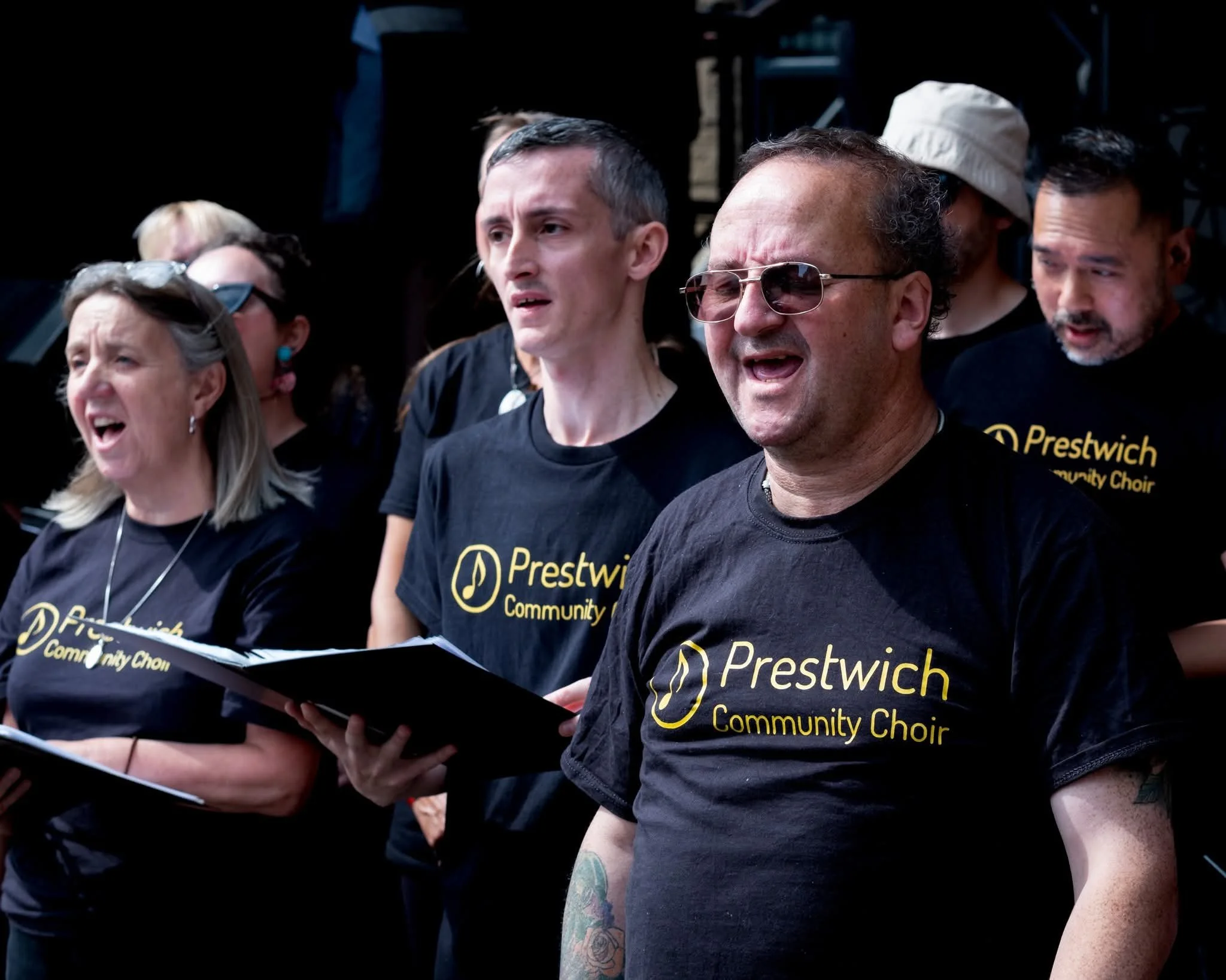 Group of people singing together, wearing black T-shirts with the text 'Prestwich Community Choir' and musical note logos, performing passionately.
