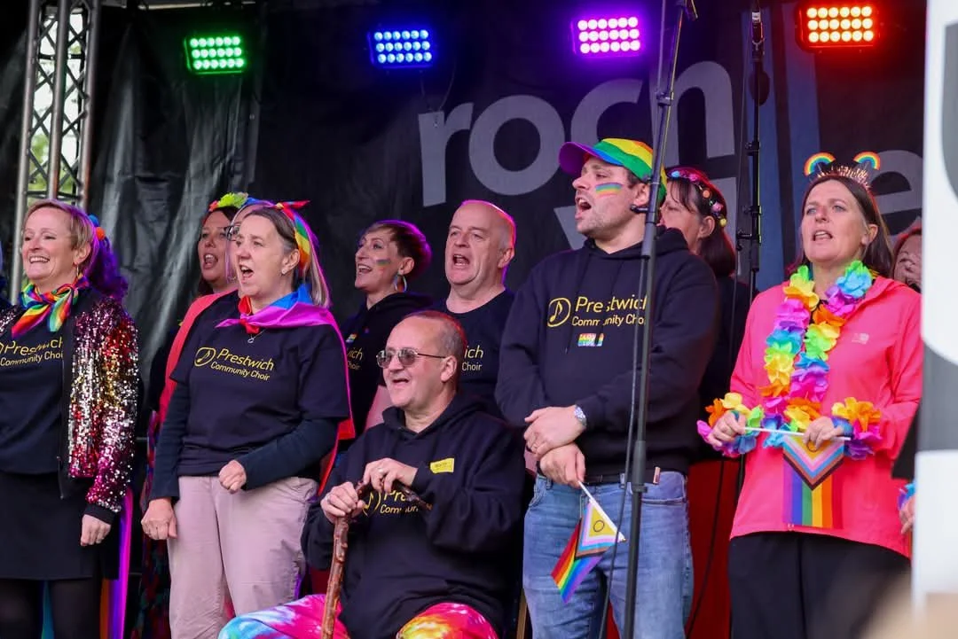 Group of people singing on stage at a pride event, wearing rainbow accessories and colorful clothing, with rainbow and community flags.