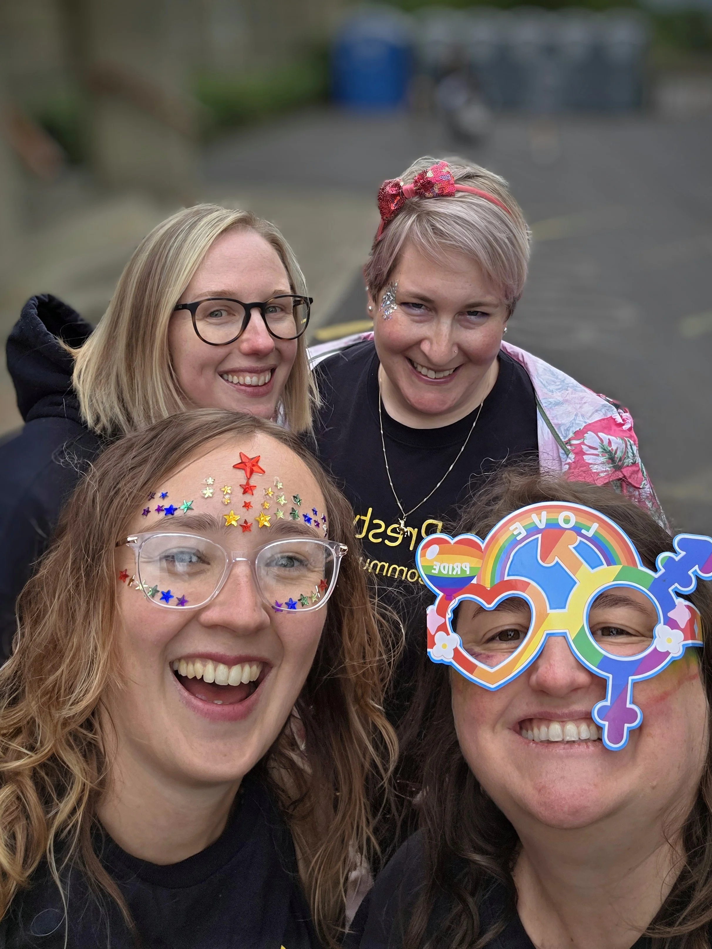 Four women smiling outdoors, with two wearing colorful star decorations on their faces and glasses, one wearing rainbow pride glasses, and another with glitter on her cheek, celebrating LGBTQ+ pride.