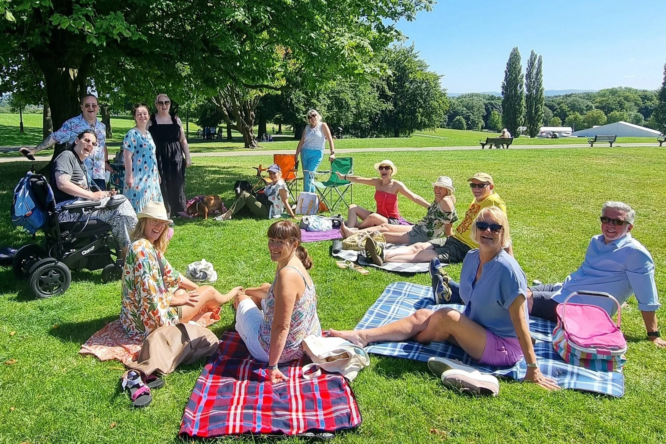 Group of people having a picnic on a sunny day in a park with green grass and trees, sitting on blankets and some with portable chairs.
