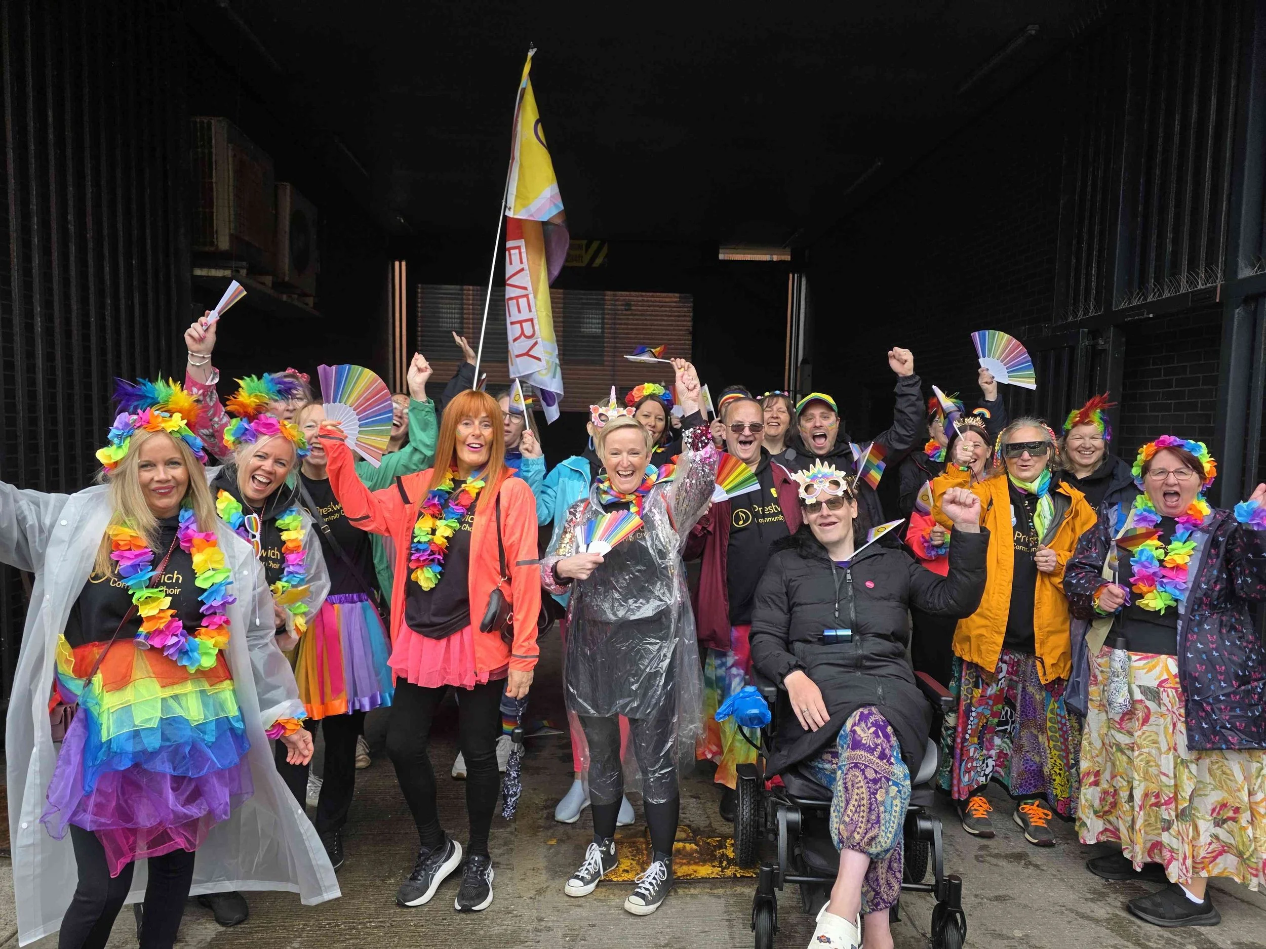 A cheerful group of choir members celebrating pride, wearing colorful rainbow-themed clothing and accessories, holding rainbow fans, and waving rainbow flags.