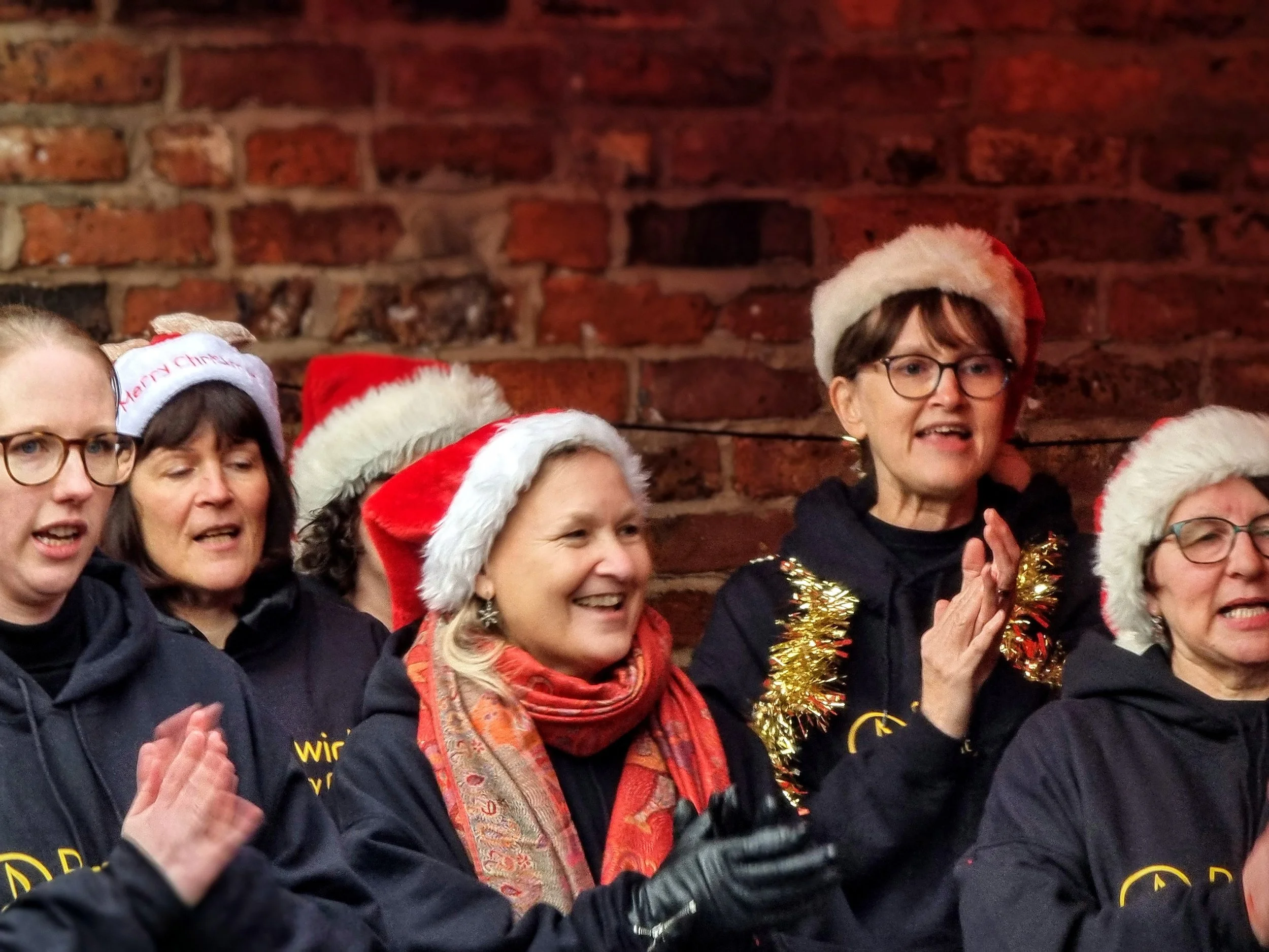 Group of women wearing Christmas hats and festive attire, singing or performing in front of a brick wall.