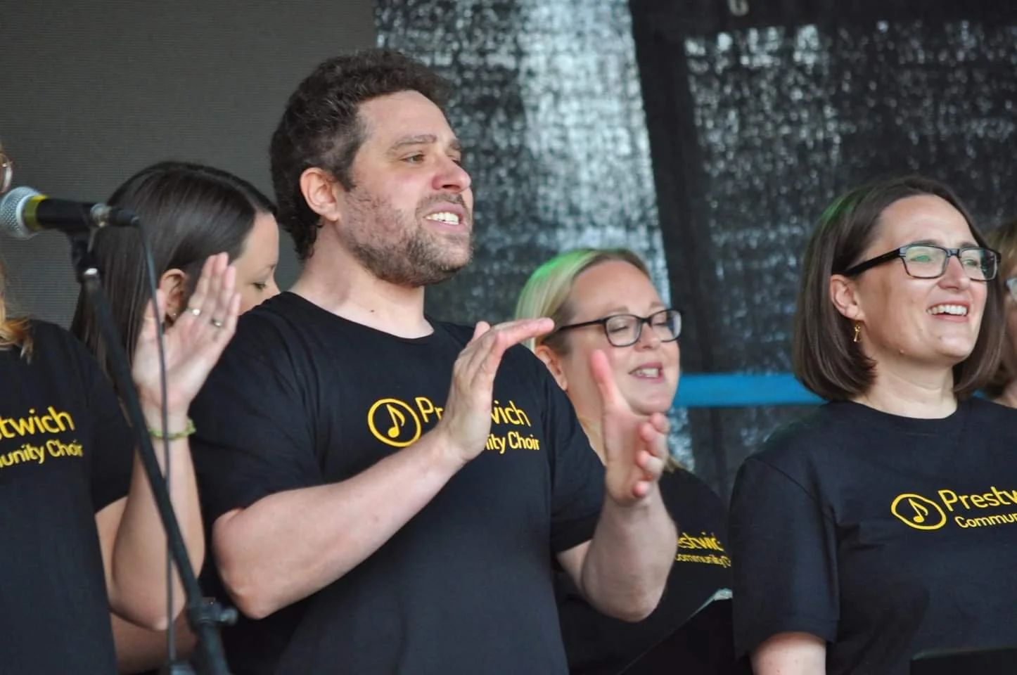 Group of people in matching black Prestwich Community Choir T-shirts, standing on stage, singing and smiling.